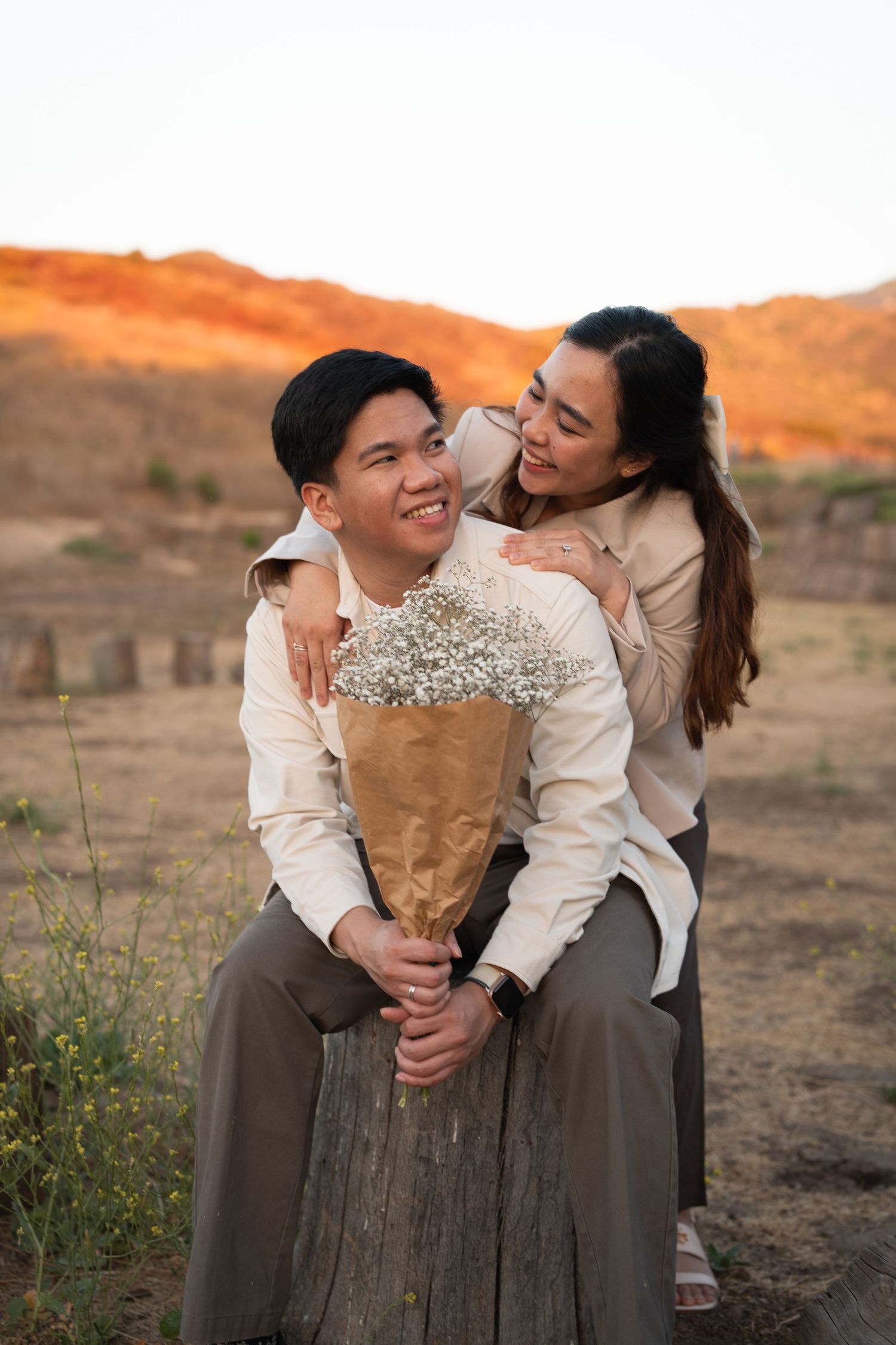 Woman embracing man from behind as he holds a bouquet of white flowers at sunset in a scenic field.