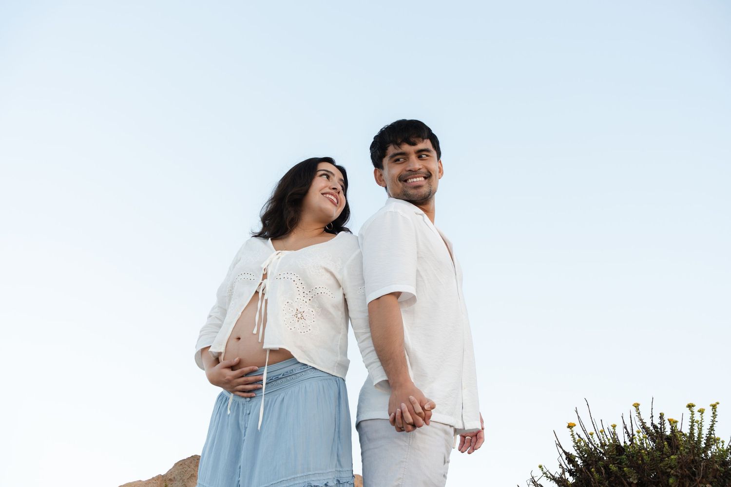 Smiling pregnant couple standing back to back against a clear blue sky near the coast.