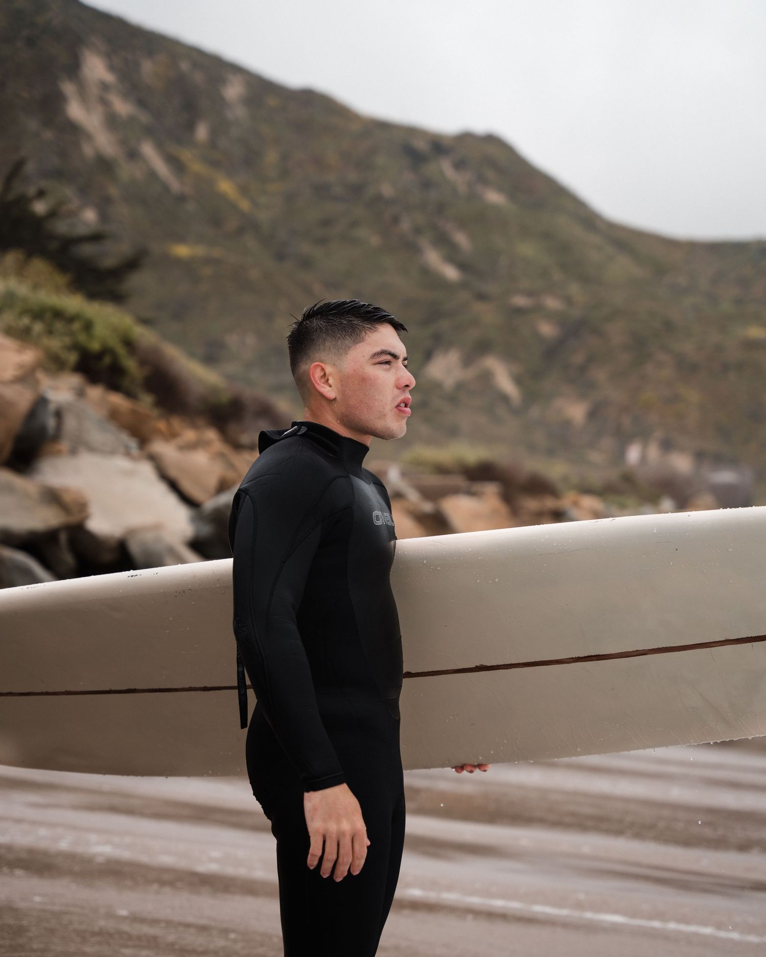 Young male surfer in black wetsuit holds white surfboard on rocky beach.