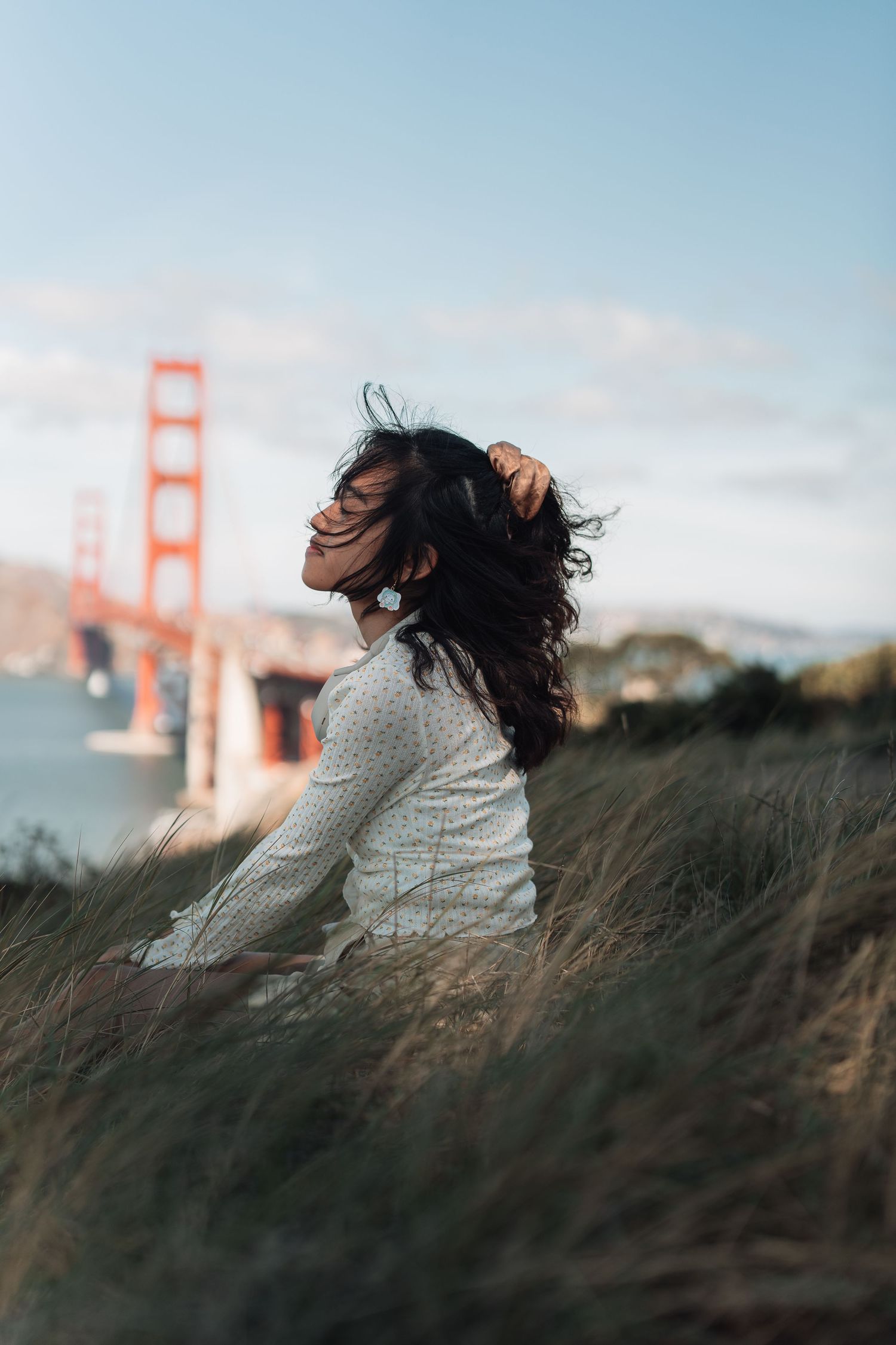 Young woman with windswept dark hair sits in grass with Golden Gate Bridge in background.