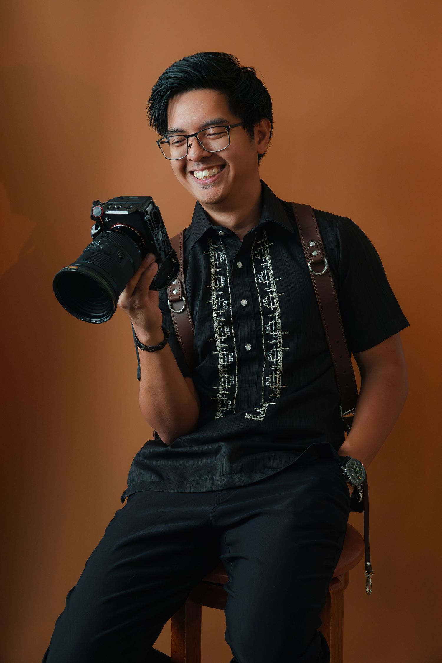 Cheerful Asian male photographer holding DSLR camera while seated, wearing black embroidered shirt with leather suspenders.