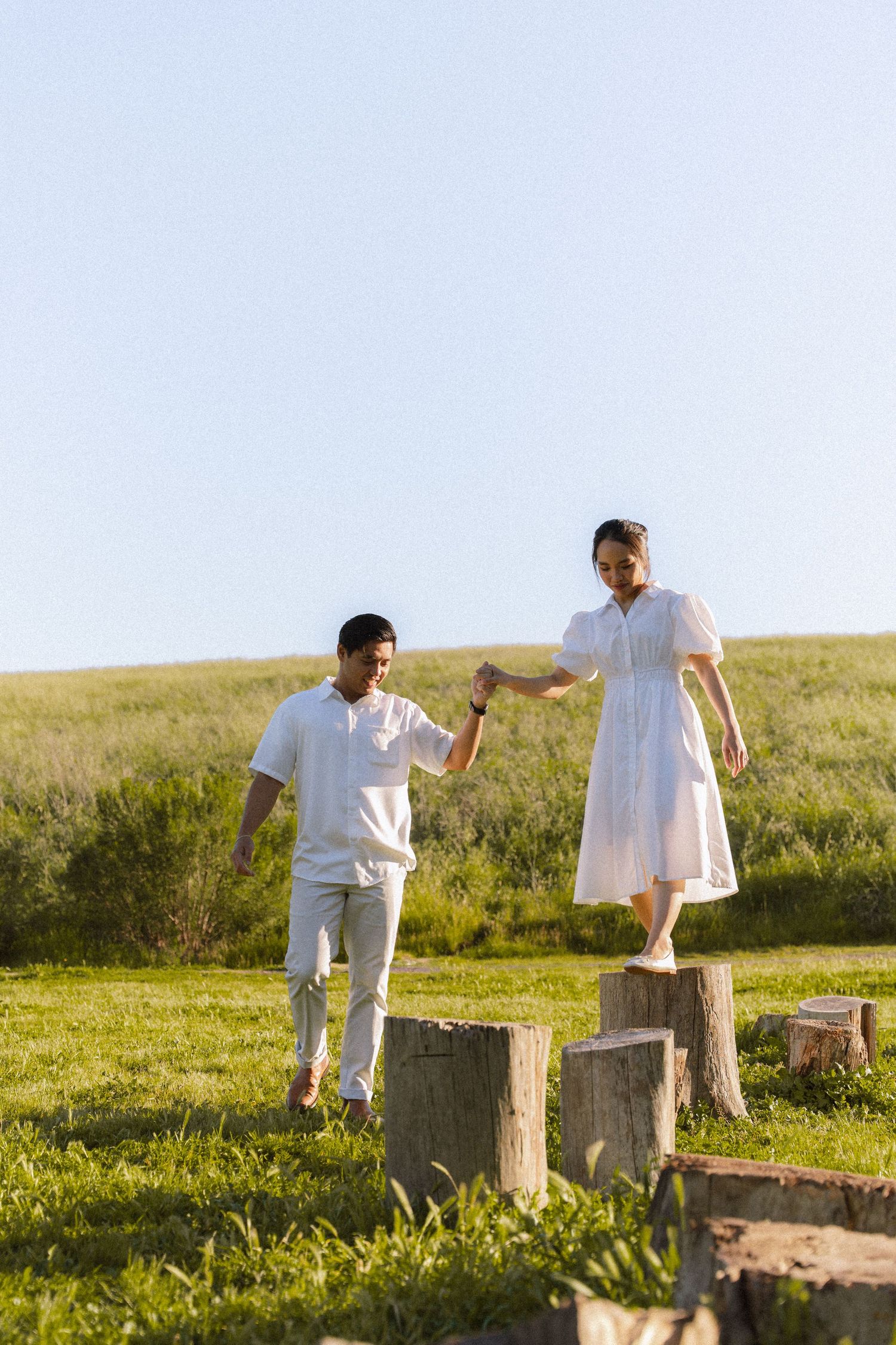 Couple in white outfits holding hands while balancing on tree stumps in a sunny green field.