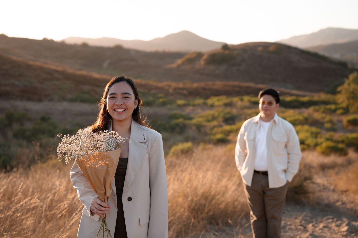 Happy woman holding a bouquet of dried flowers while her partner stands in the background at golden hour.