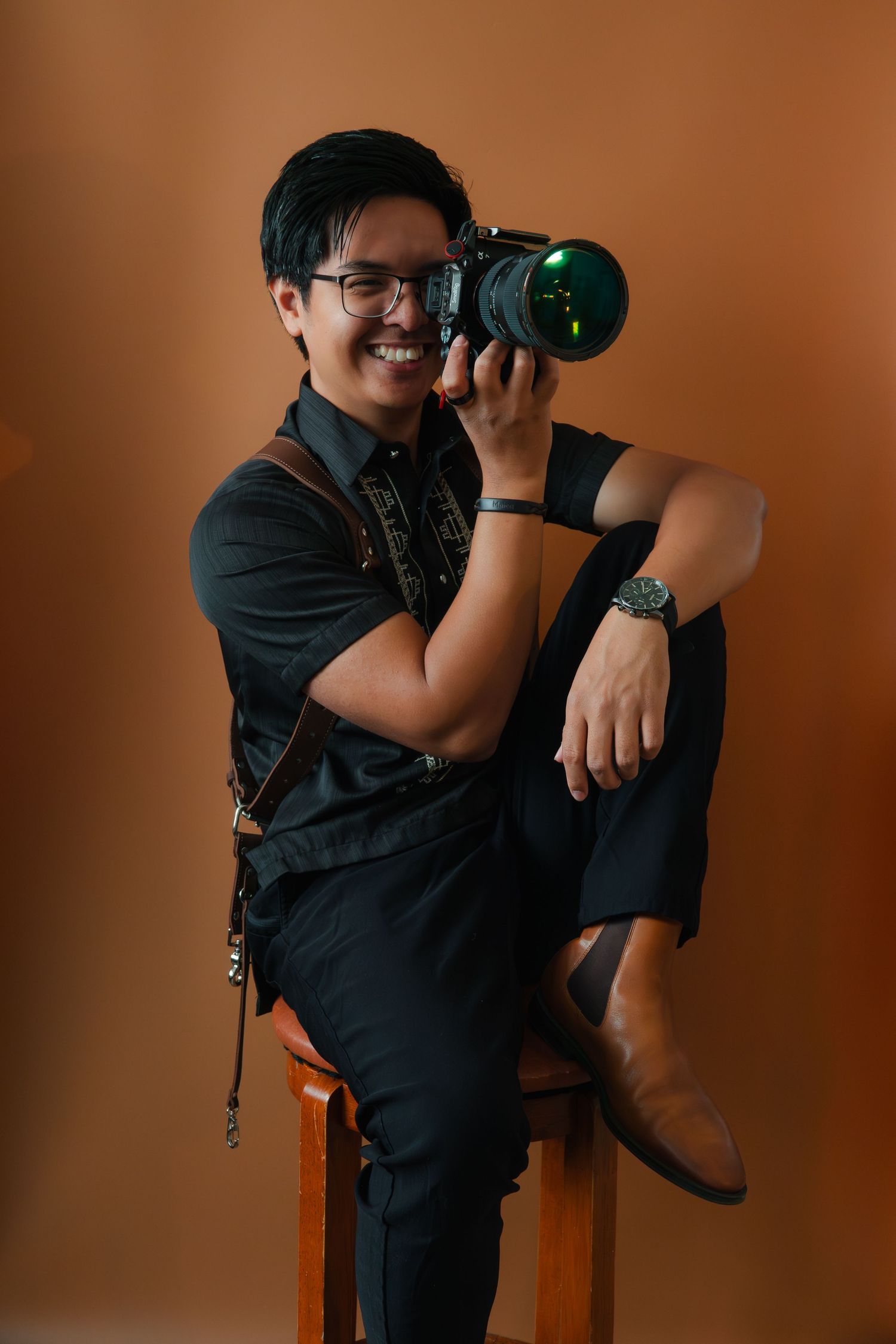 Smiling photographer in black outfit posing with DSLR camera on wooden stool.