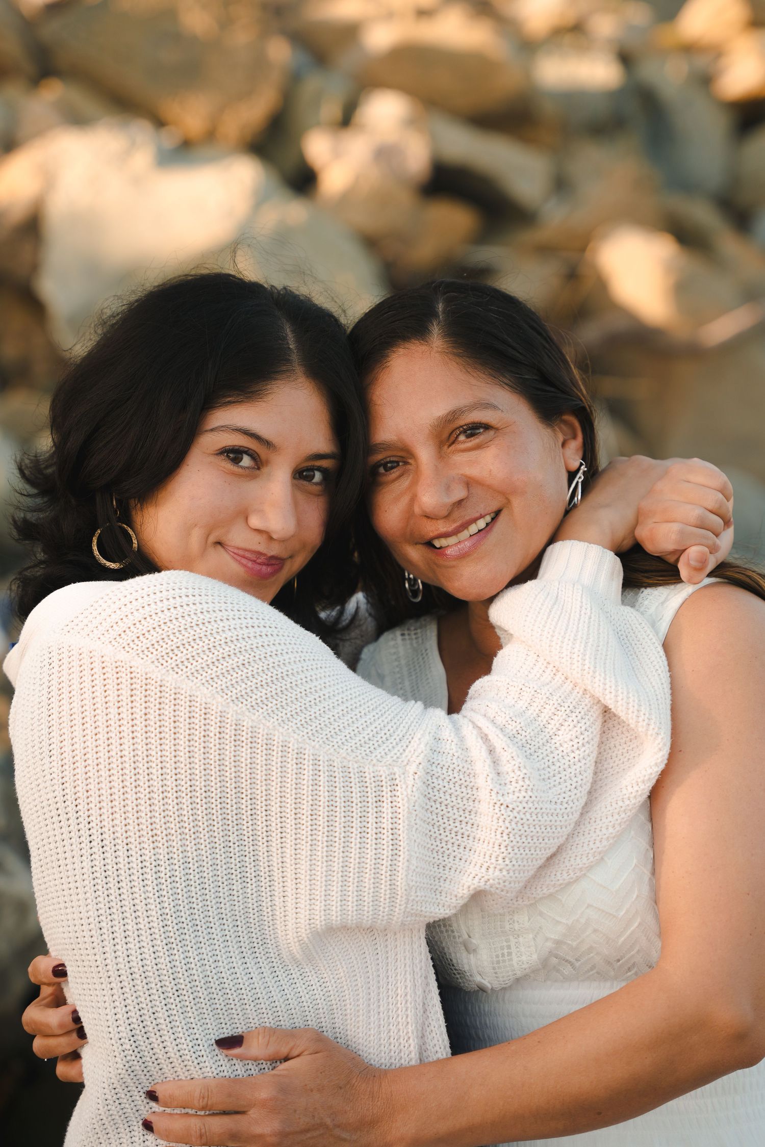 Two women embracing and smiling warmly in front of rocky background at sunset.