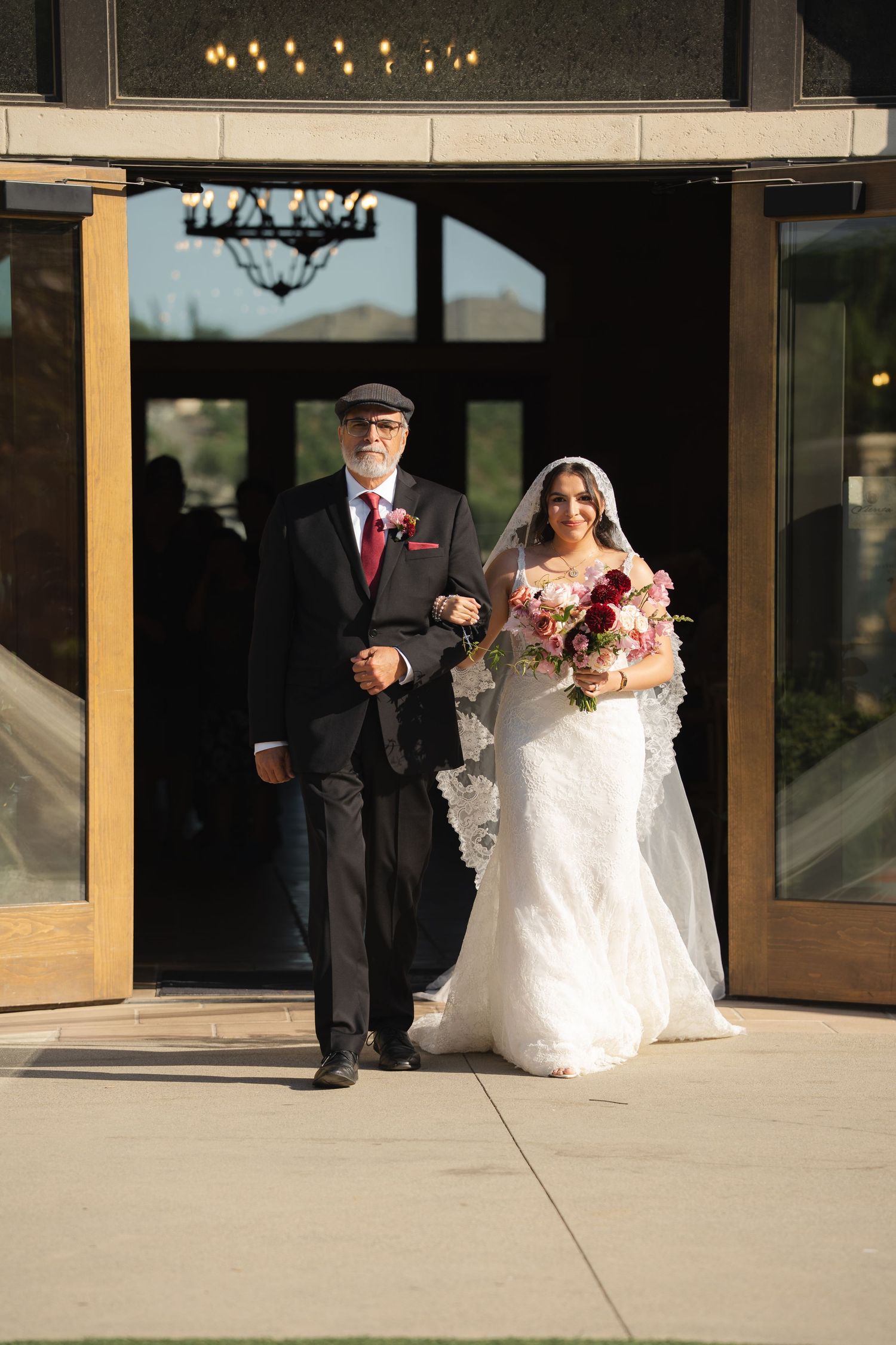 Bride in lace mermaid gown with red bouquet being escorted by father through venue entrance doors.