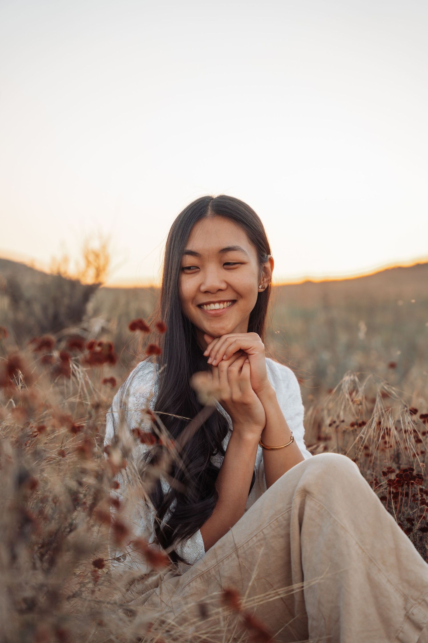 Smiling young Asian woman sitting in golden field at sunset with hands clasped together.