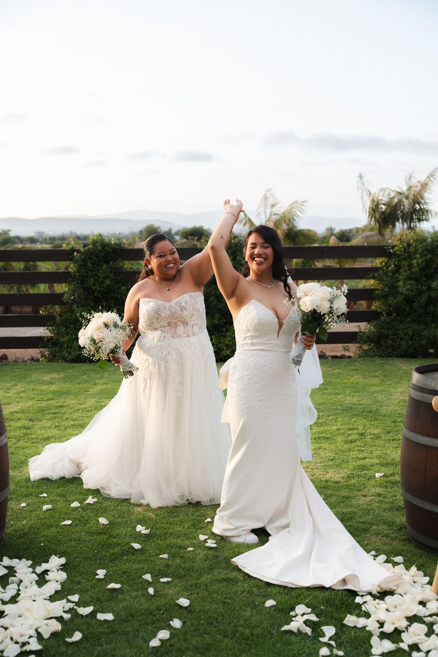 Two brides celebrate joyfully on a rose petal-lined lawn holding white bouquets after their ceremony.