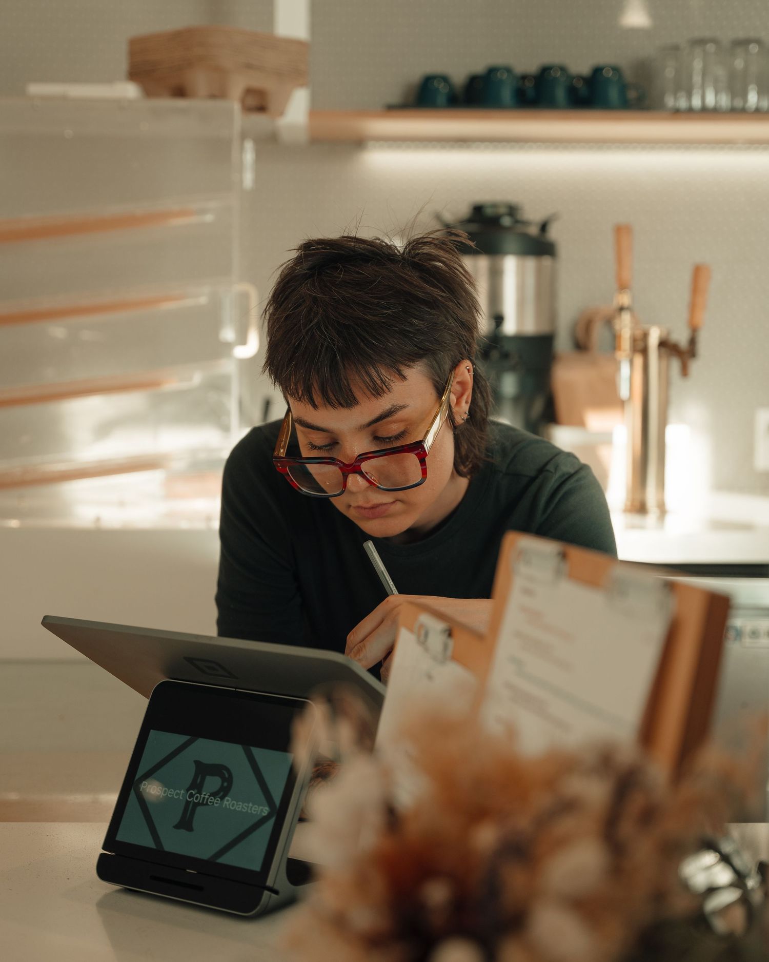 Barista with red glasses writing on notepad beside tablet POS system at modern coffee shop counter.