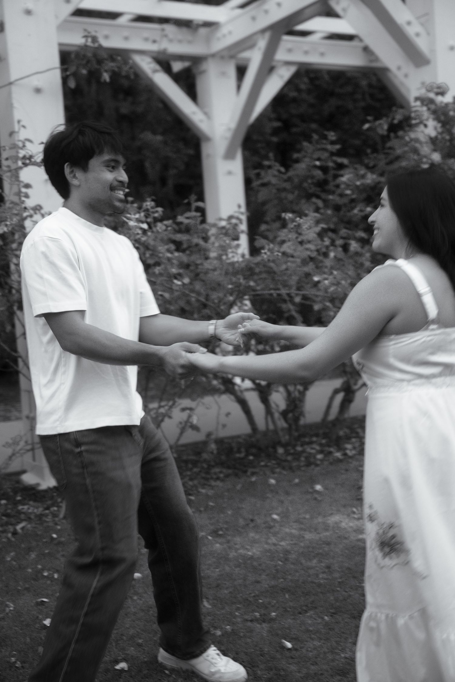 Black and white photo of a smiling couple holding hands in a garden with pergola.