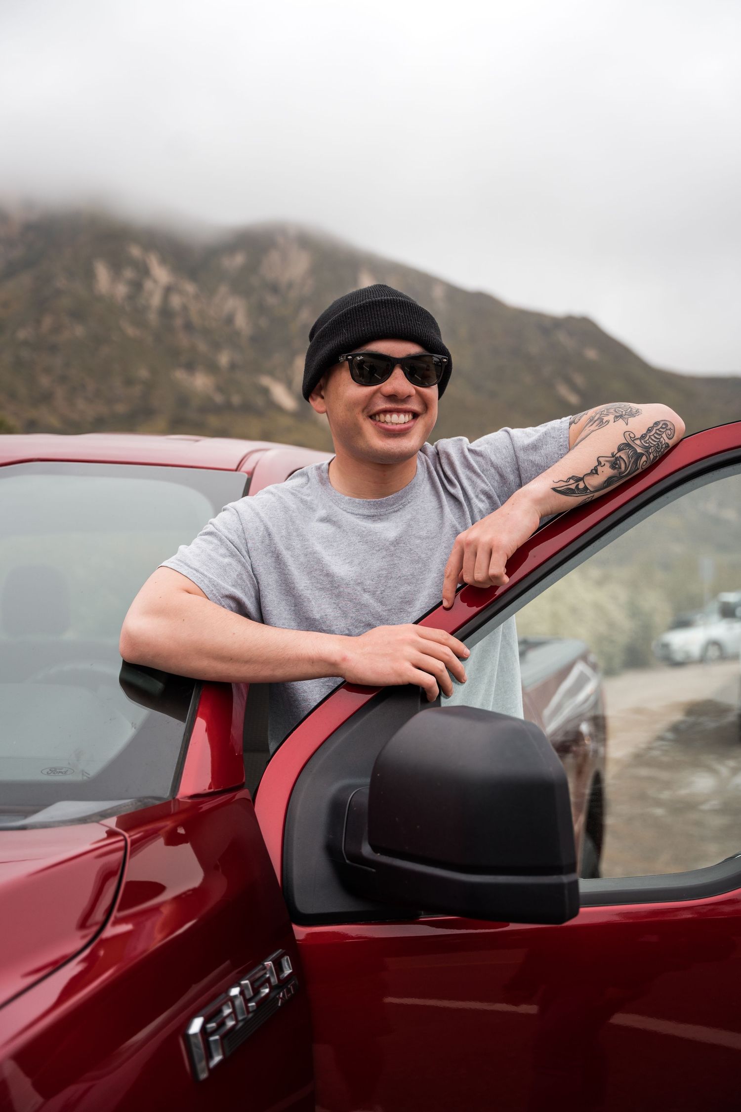 Smiling man in gray tee and black beanie leans on red pickup truck in mountains.