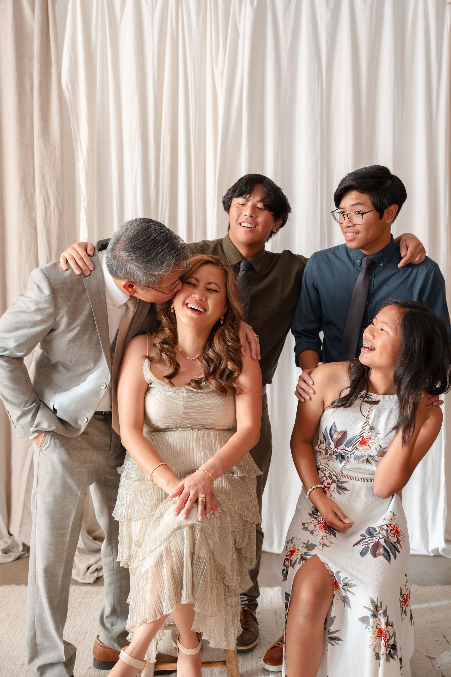 Happy Asian family of five posing together, laughing and embracing in front of white curtain backdrop.