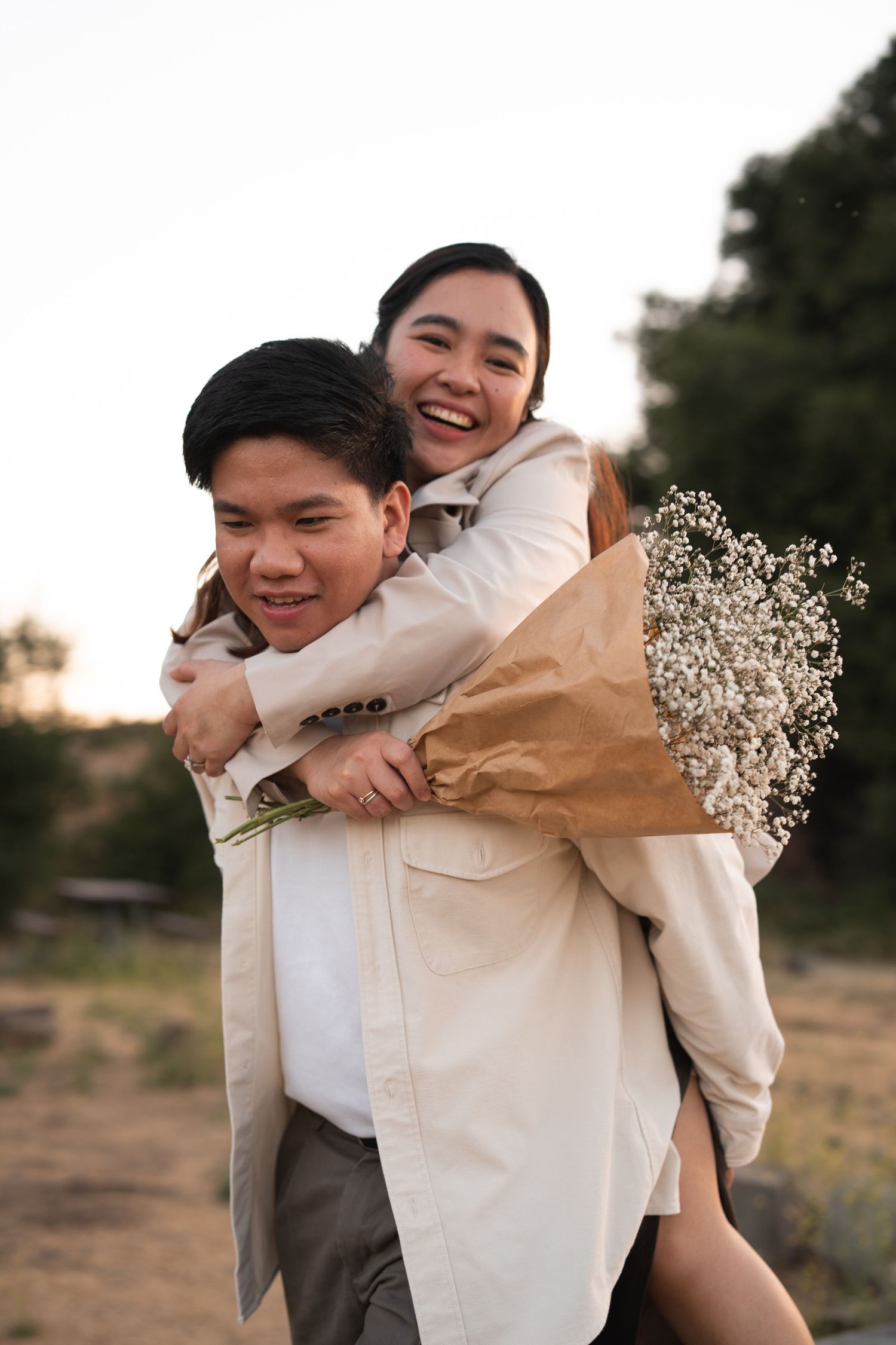 Happy couple laughing as man gives woman a piggyback ride while she holds flowers.