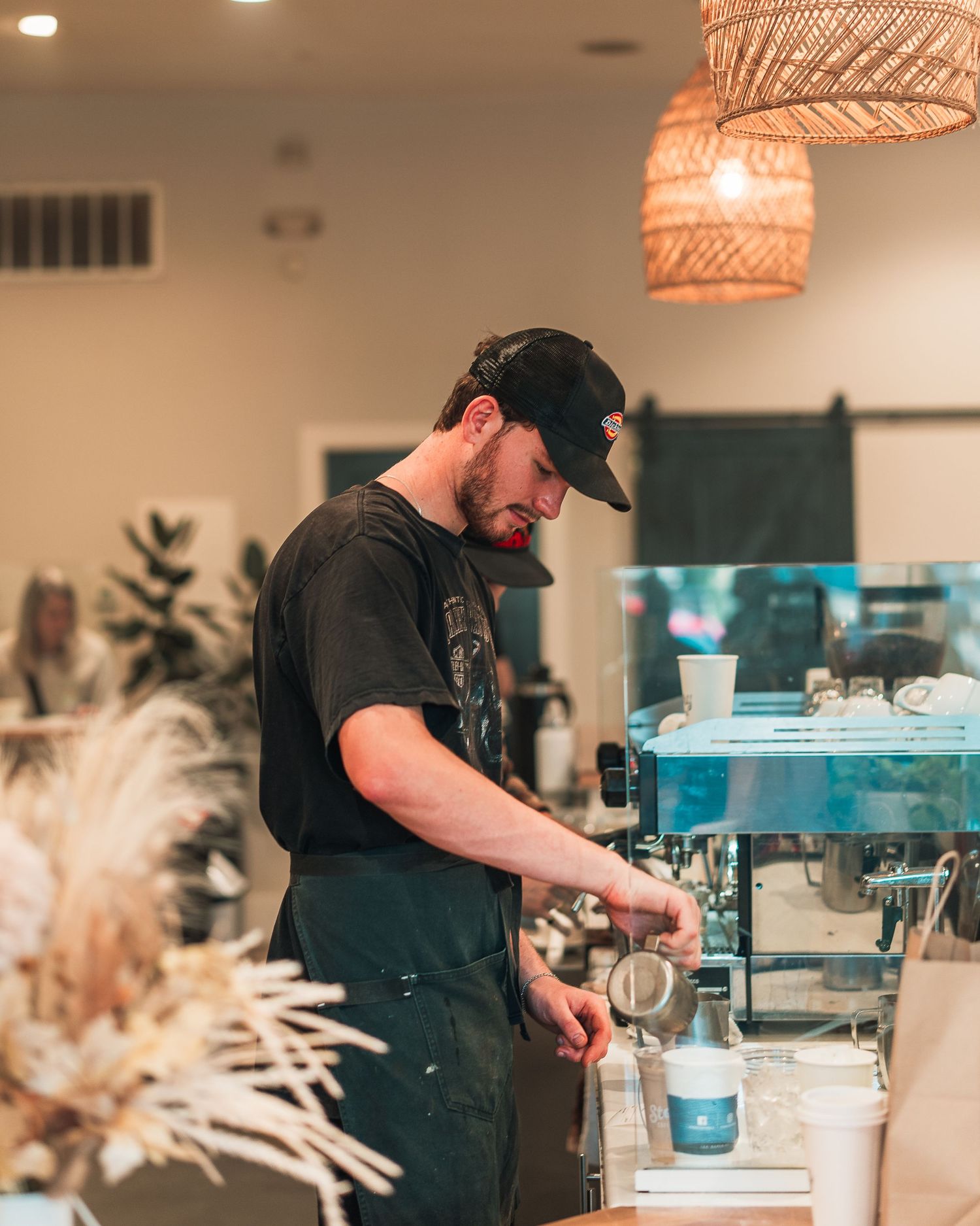 Barista in black cap and apron preparing espresso at a coffee machine in a bright cafe.