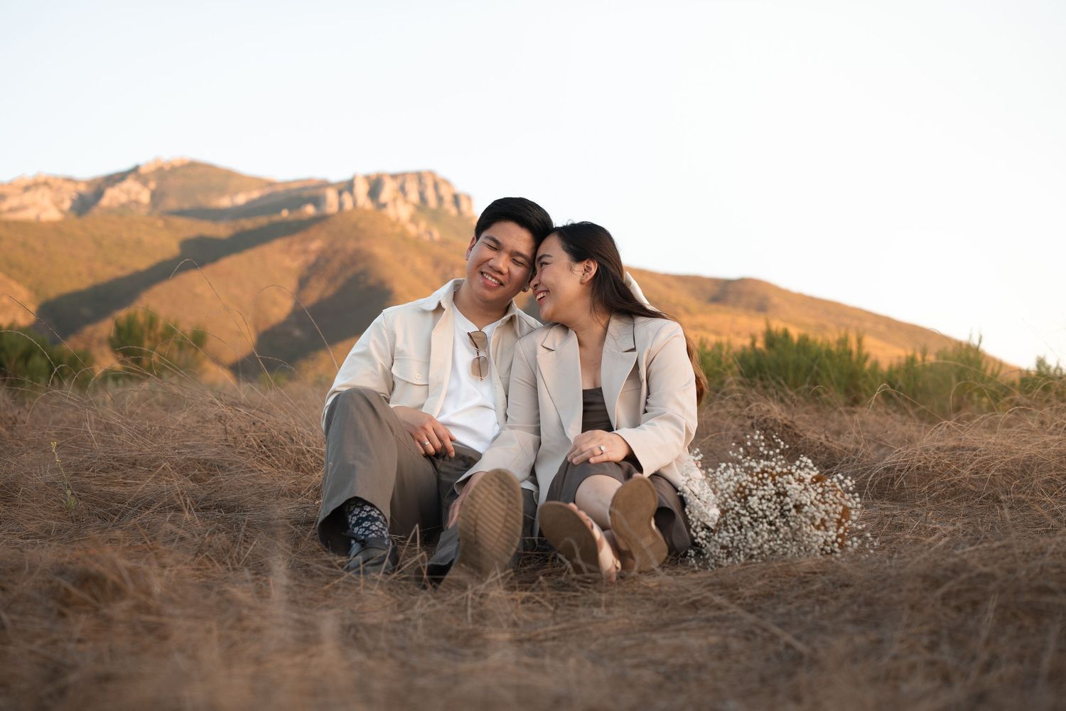 Happy couple sitting together in a golden field with mountains lit by warm sunset light behind them.