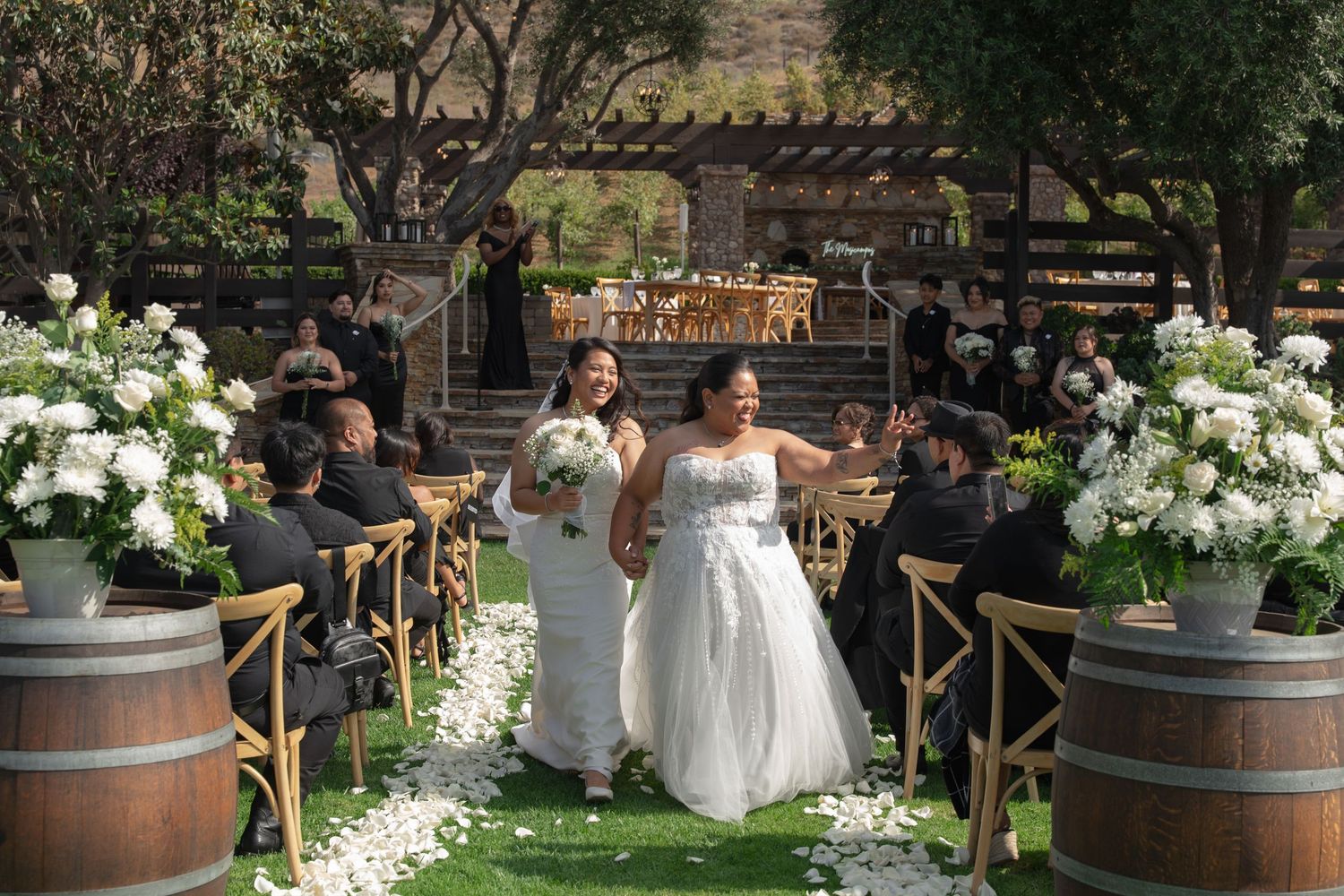 Two brides in white gowns celebrating joyfully while walking down aisle lined with white florals.