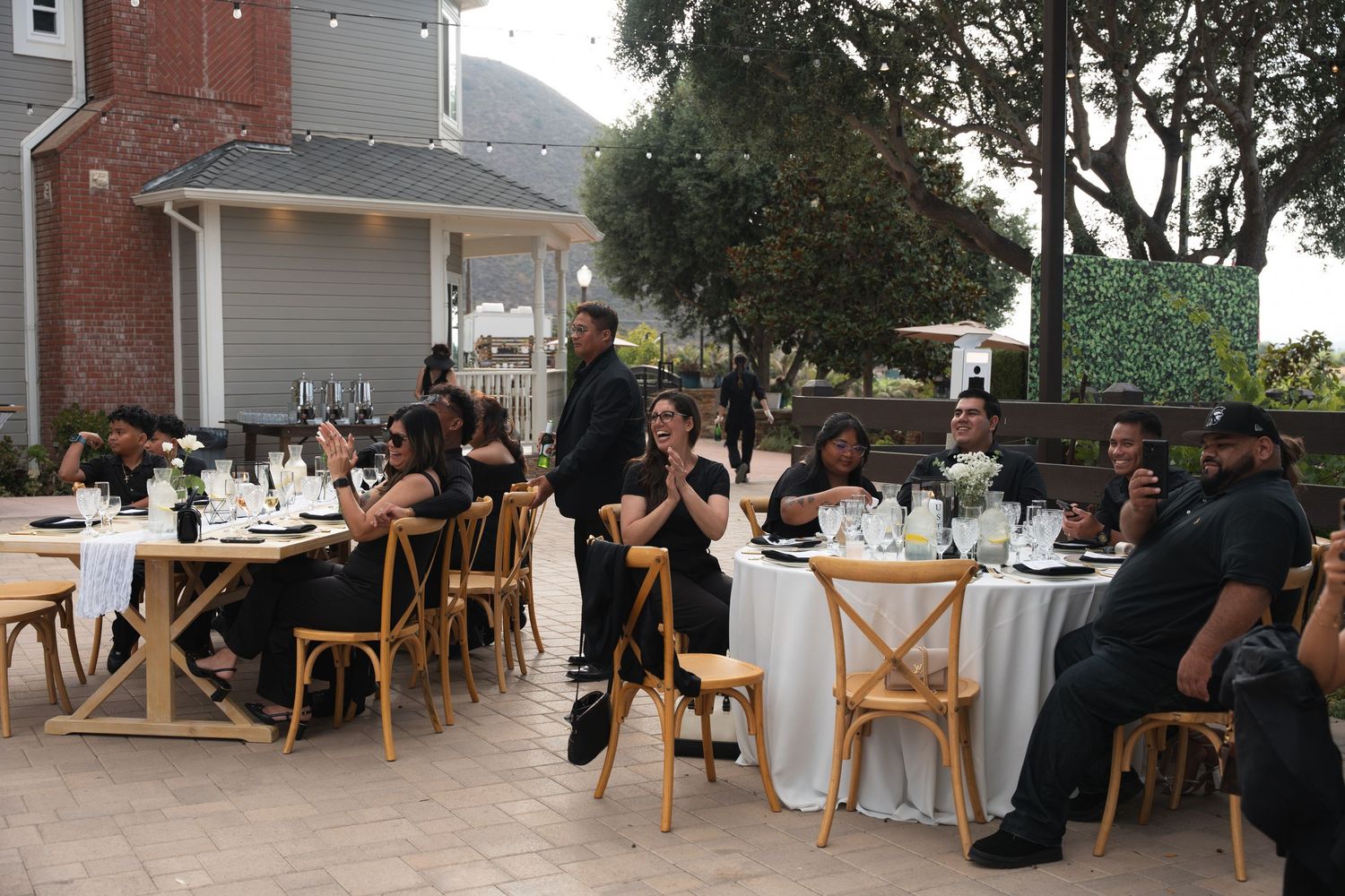 Wedding guests in black attire seated at rustic wooden tables during outdoor reception.