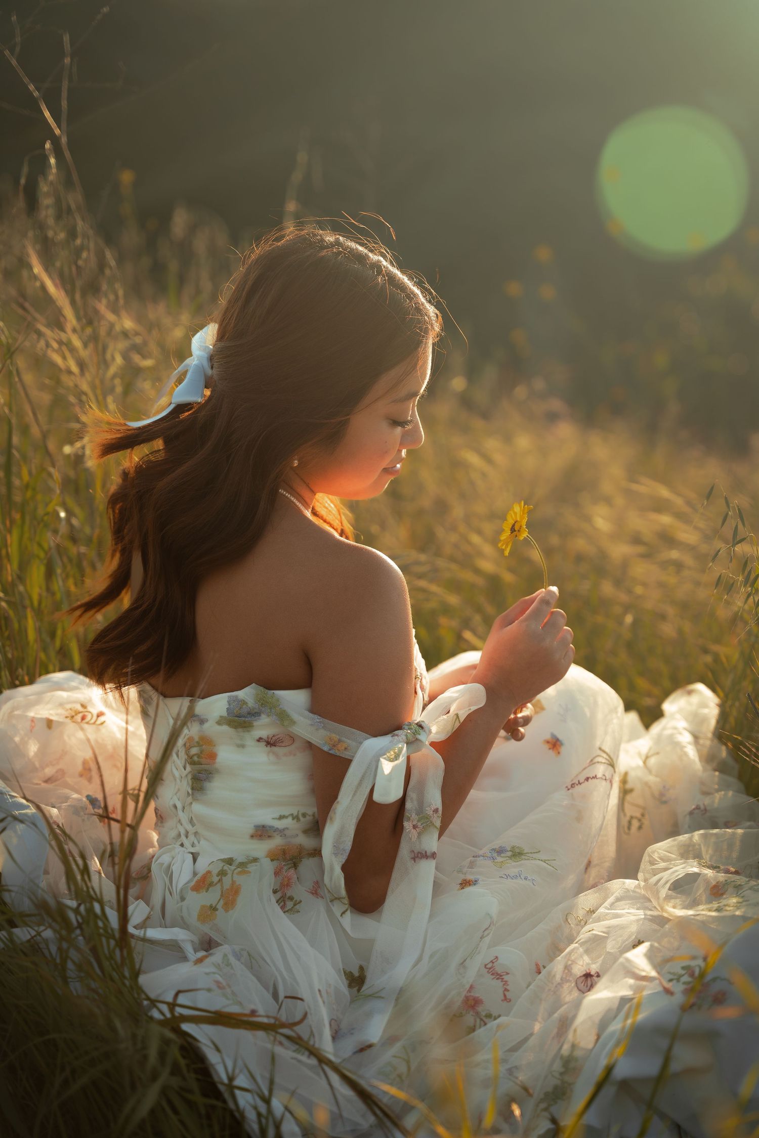 A young woman in a floral dress sits in a sunlit field holding a wildflower with golden lens flare.