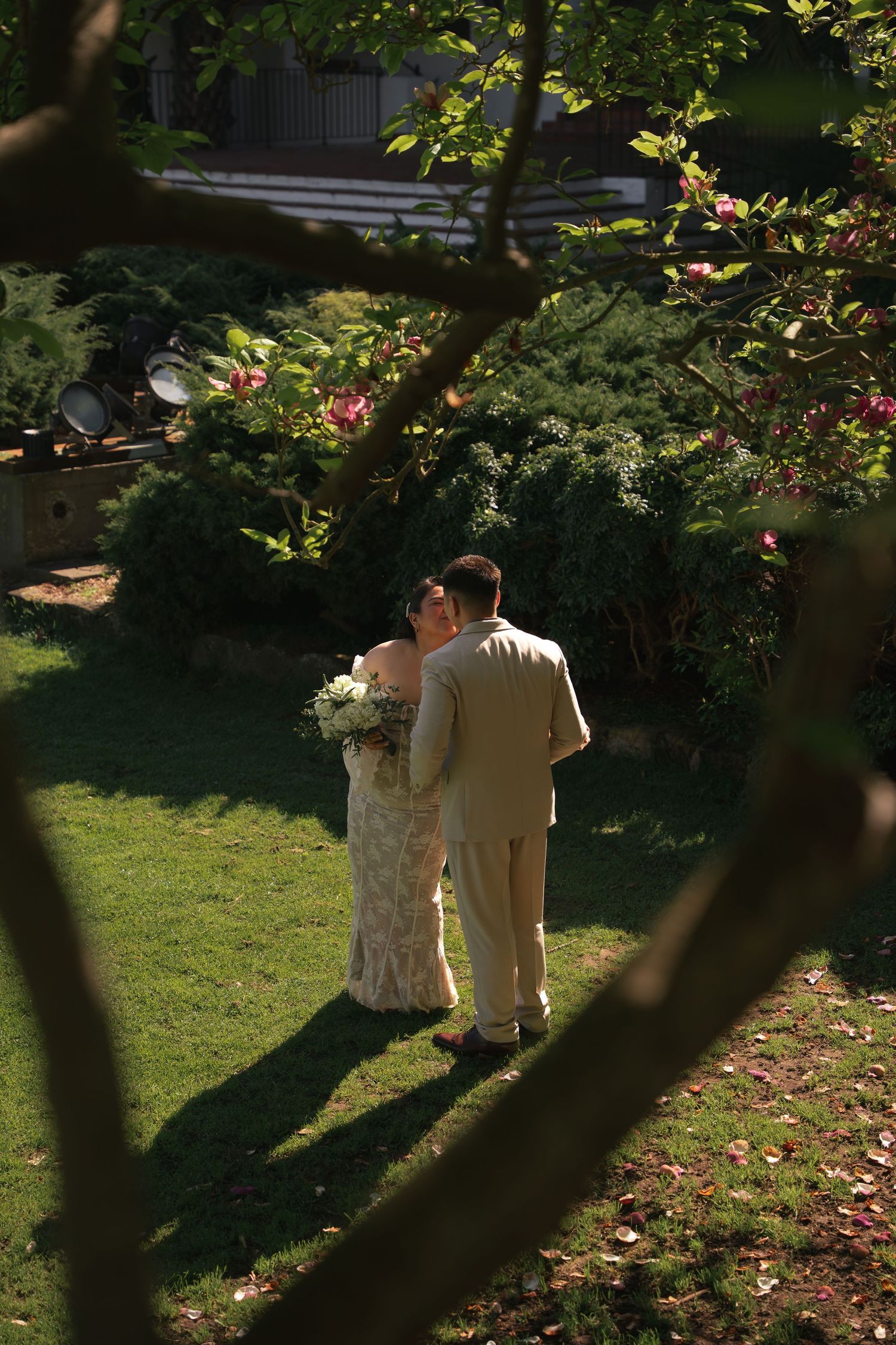 Bride and groom sharing intimate moment on lush garden lawn framed by flowering trees.