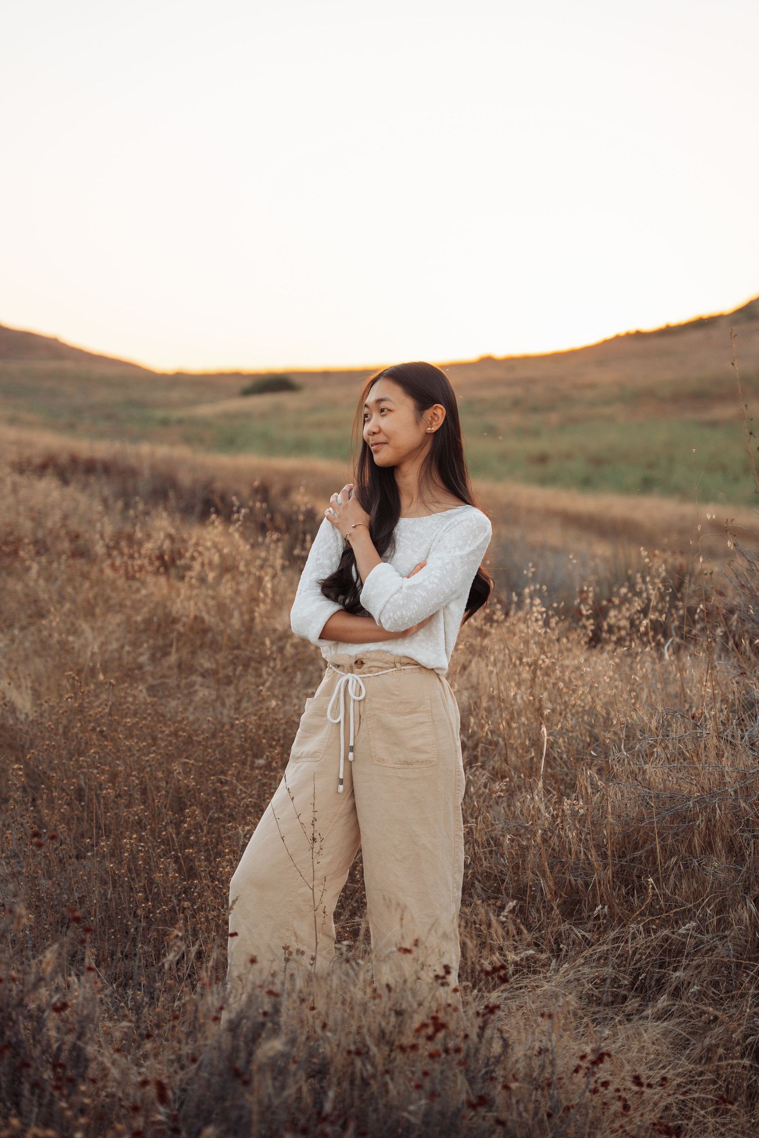 Young woman in white top and beige wide-leg pants standing in a golden field at sunset.