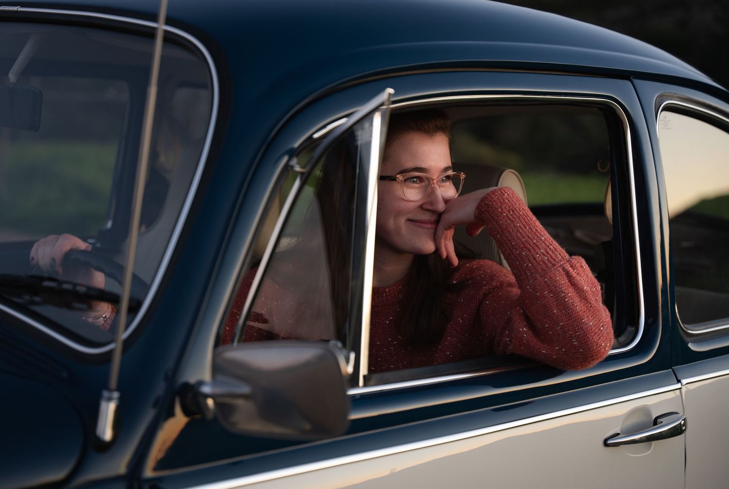 Smiling woman in red sweater leaning on vintage car window in warm golden light.