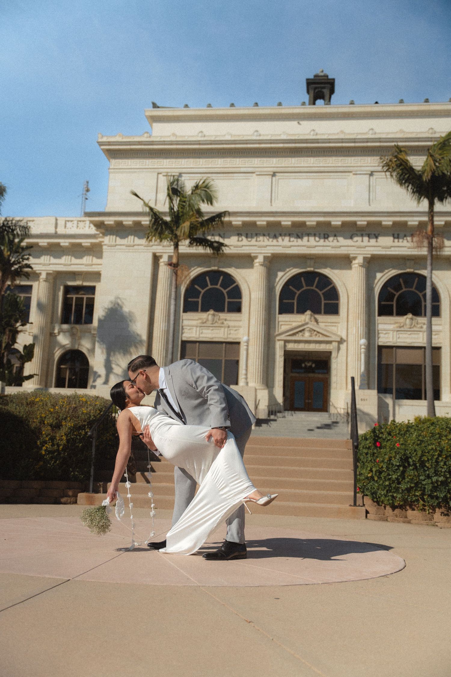 Groom dips bride in white dress for a kiss in front of historic city hall building.