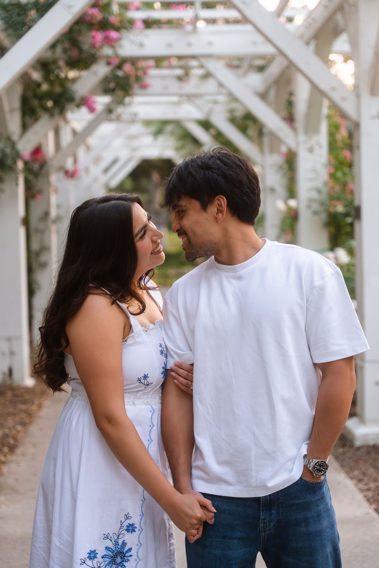 Couple gazing at each other under a white floral pergola archway in a garden.