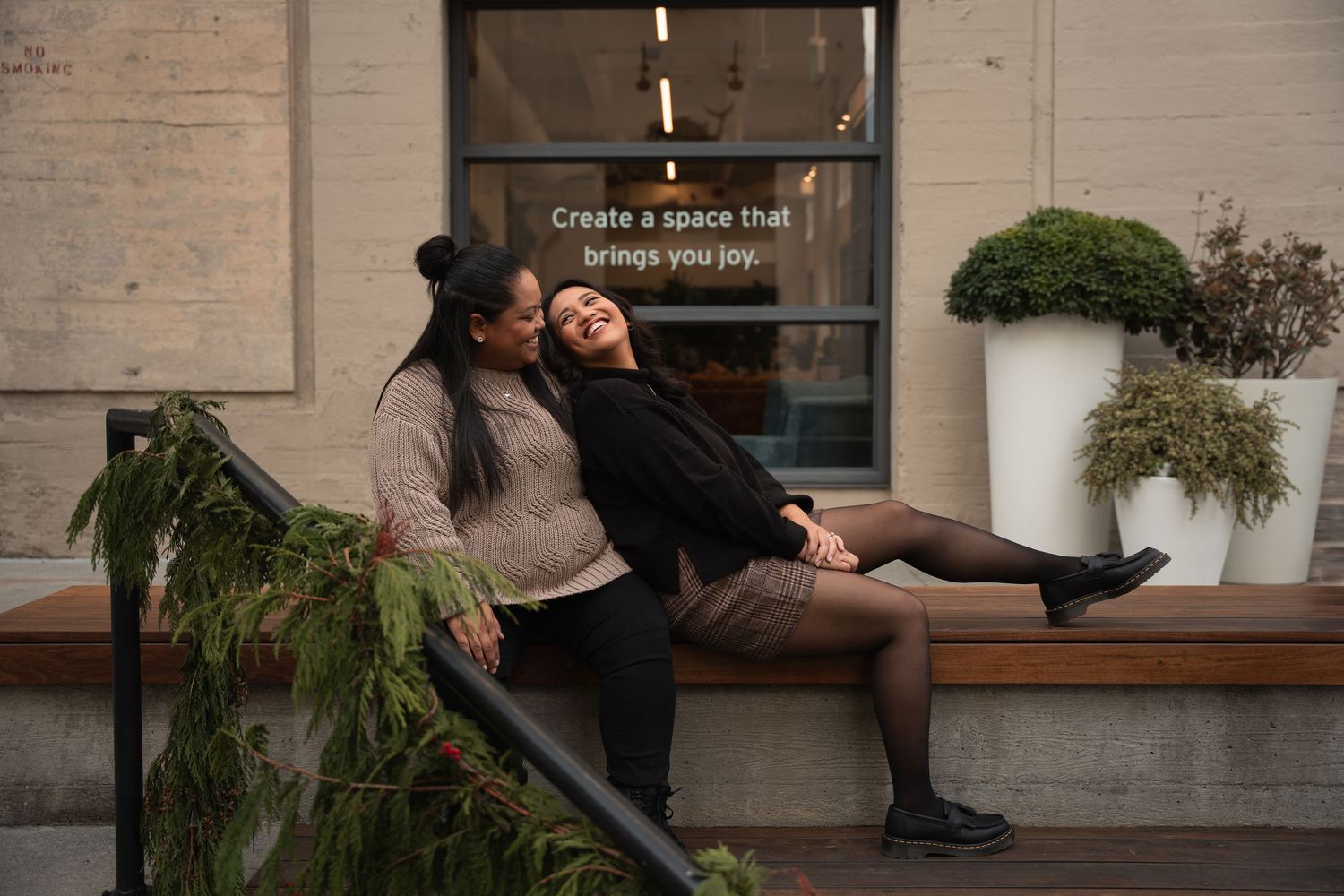 Two women sitting on an outdoor bench laughing together near a sign that reads create a space that brings you joy.