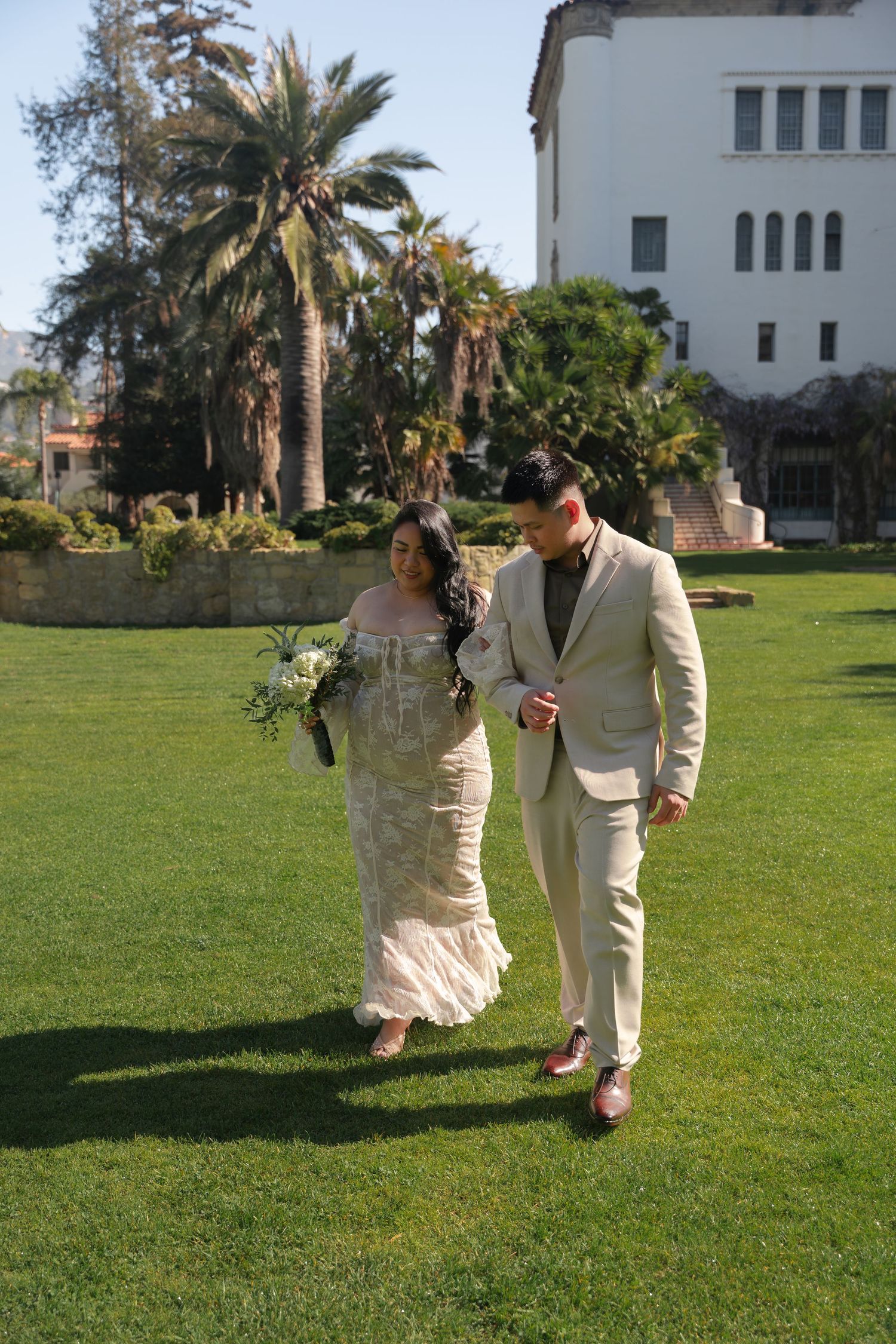 Bride and groom walking on sunny lawn with palm trees outside a white wedding venue.