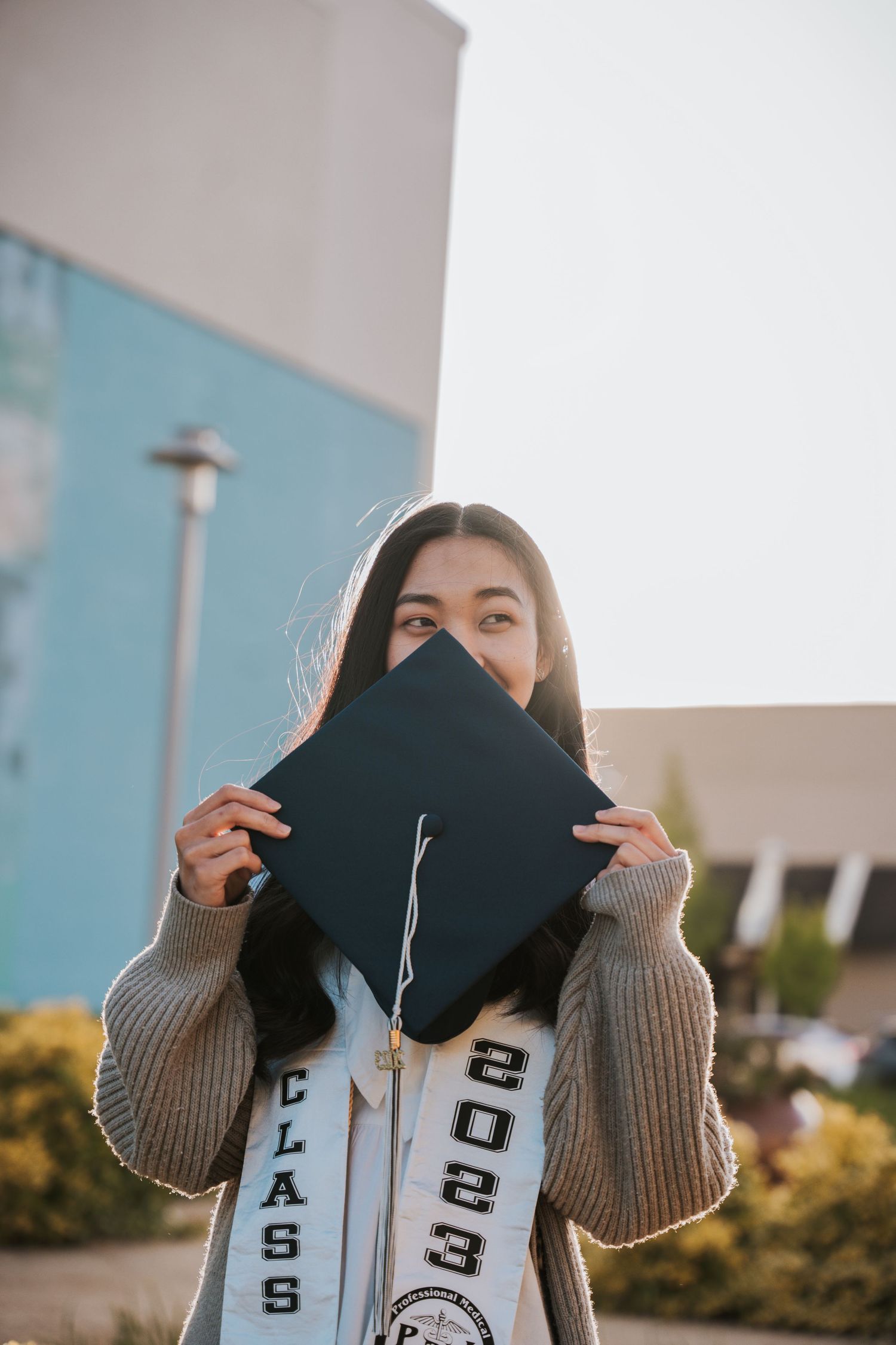 Graduate holding black mortarboard cap over face wearing Class of 2023 stole outdoors.