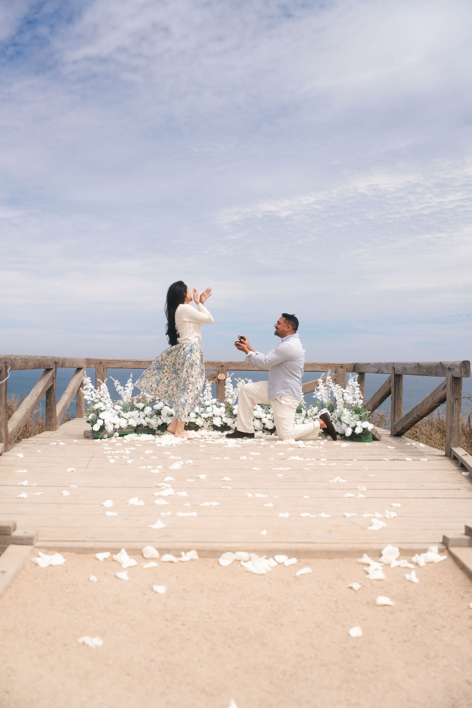 Man proposing on one knee to woman on sandy deck surrounded by white flowers and petals.