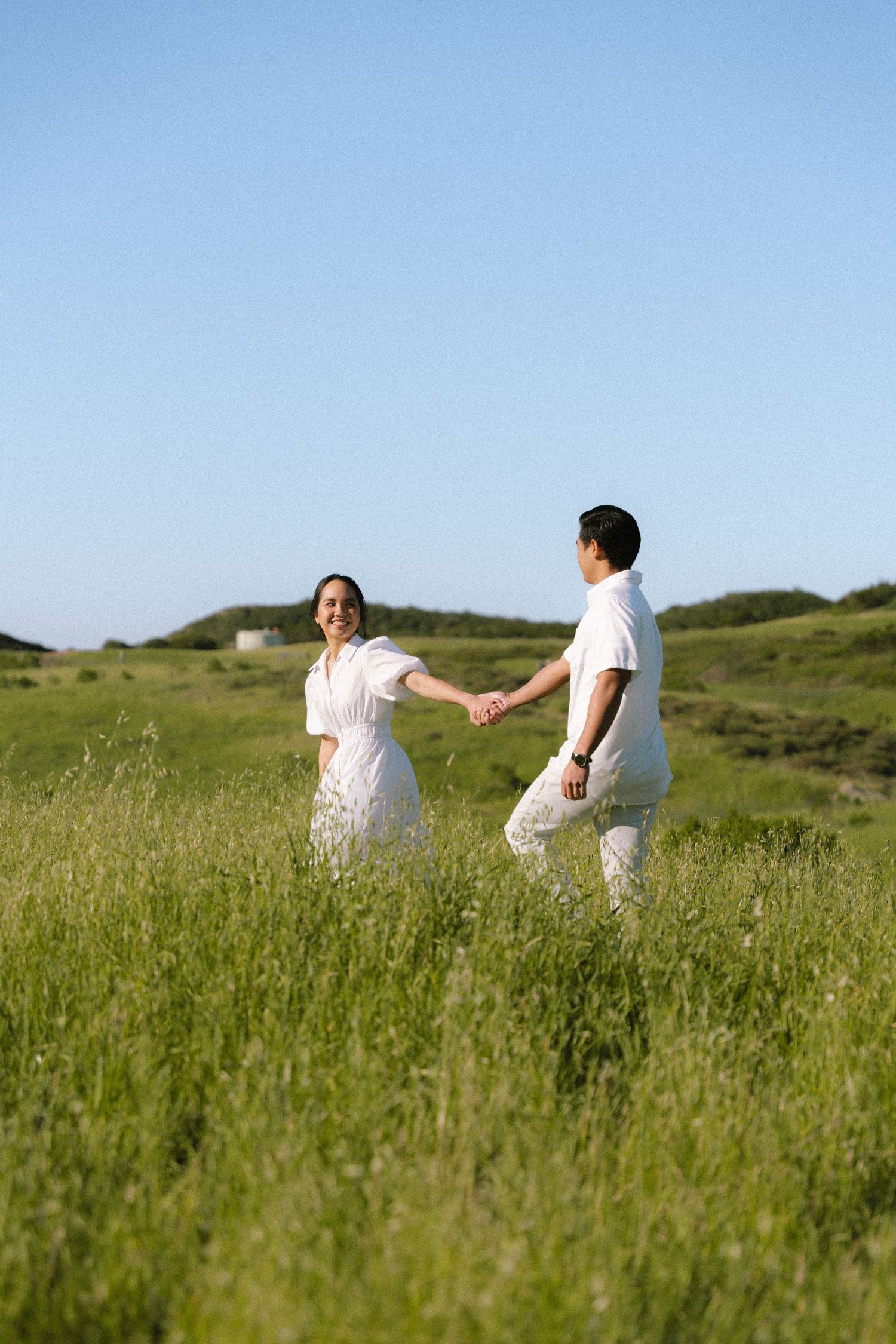 Happy couple in white holding hands walking through tall green grass under clear blue sky.