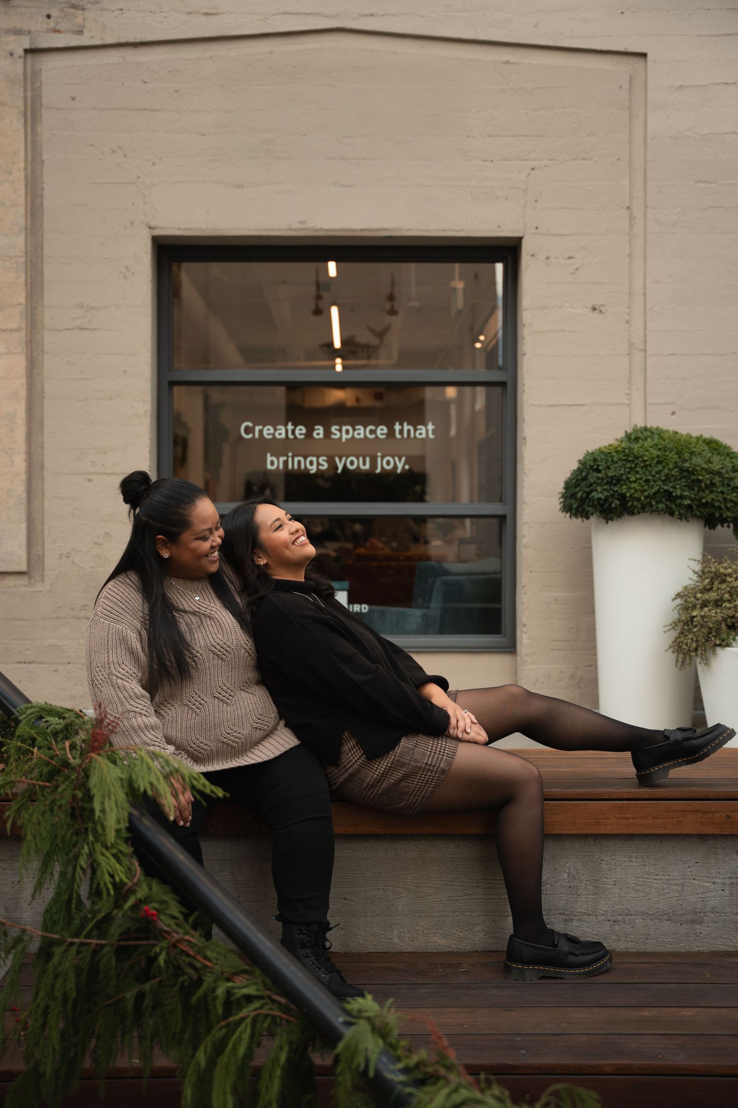 Two women laugh and sit together on outdoor steps near a window reading Create a space that brings you joy.