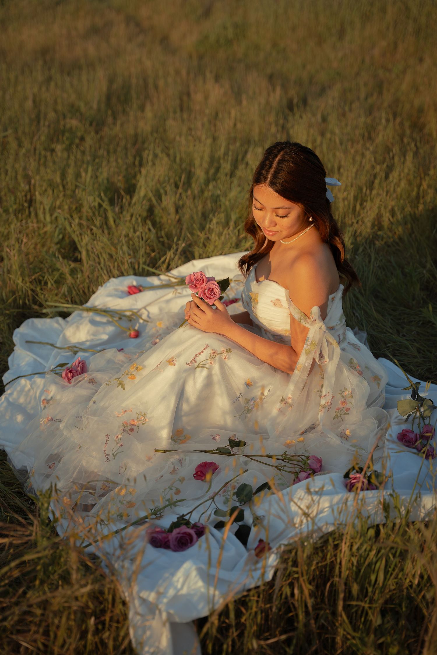 Young woman in white floral gown sitting in golden field holding pink roses at sunset.
