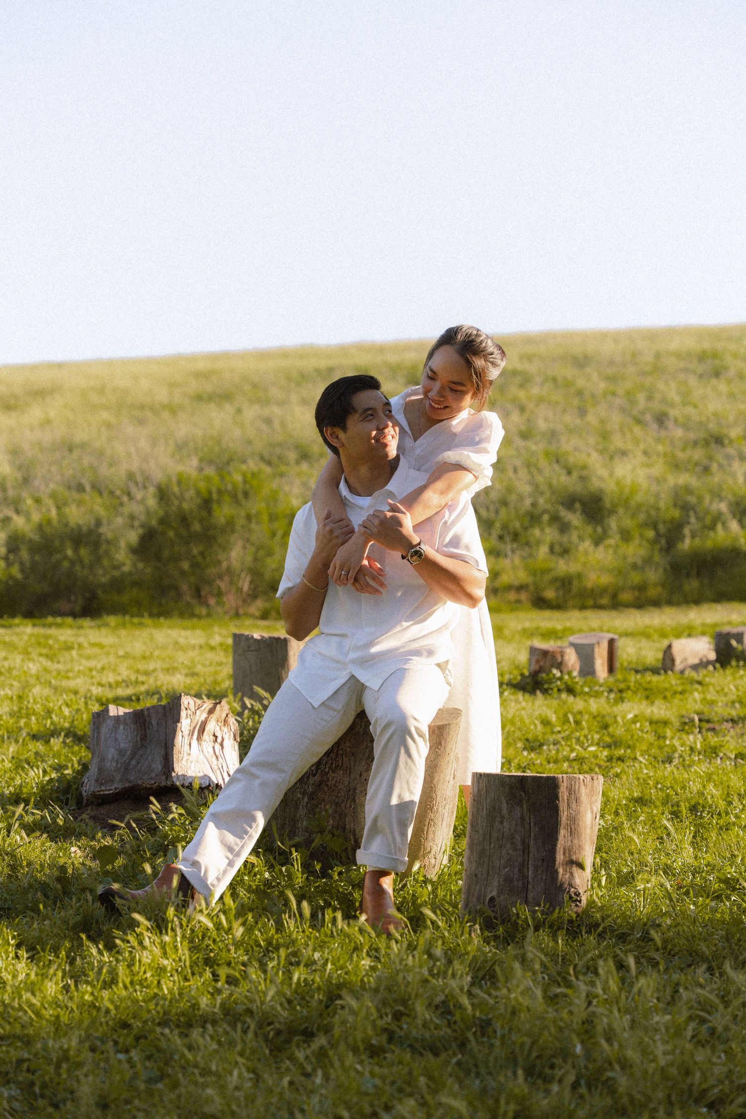 Happy couple in white outfits embracing near rustic wooden stumps on sunny green hillside.