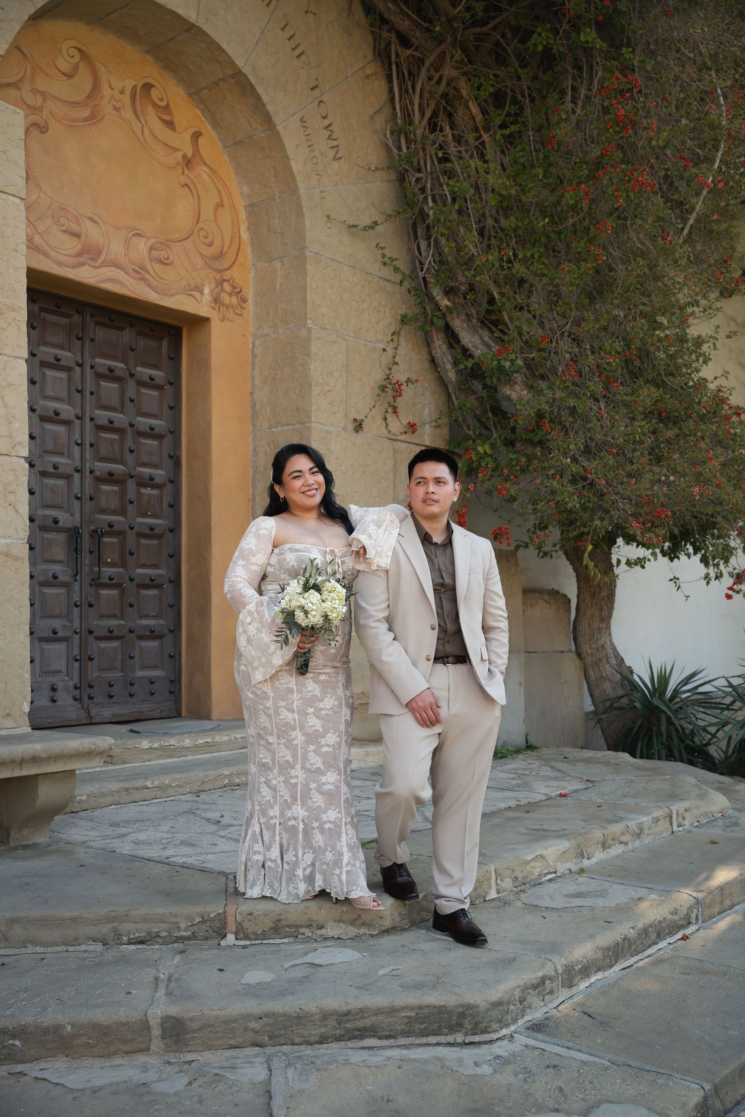 Bride in lace gown and groom in beige suit posing on church steps with bouquet of white flowers.