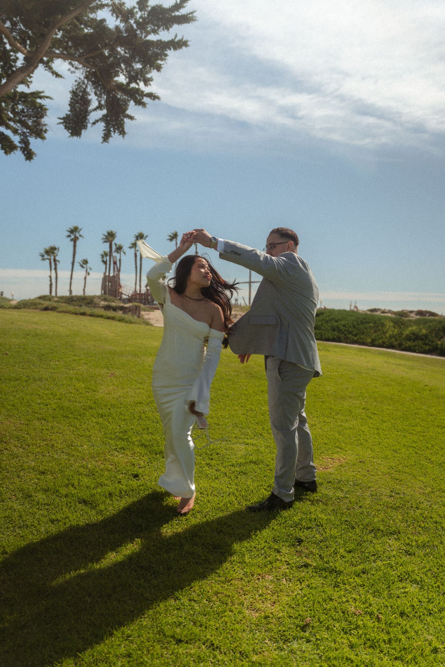 Bride and groom dance joyfully on green lawn with palm trees and ocean in background.