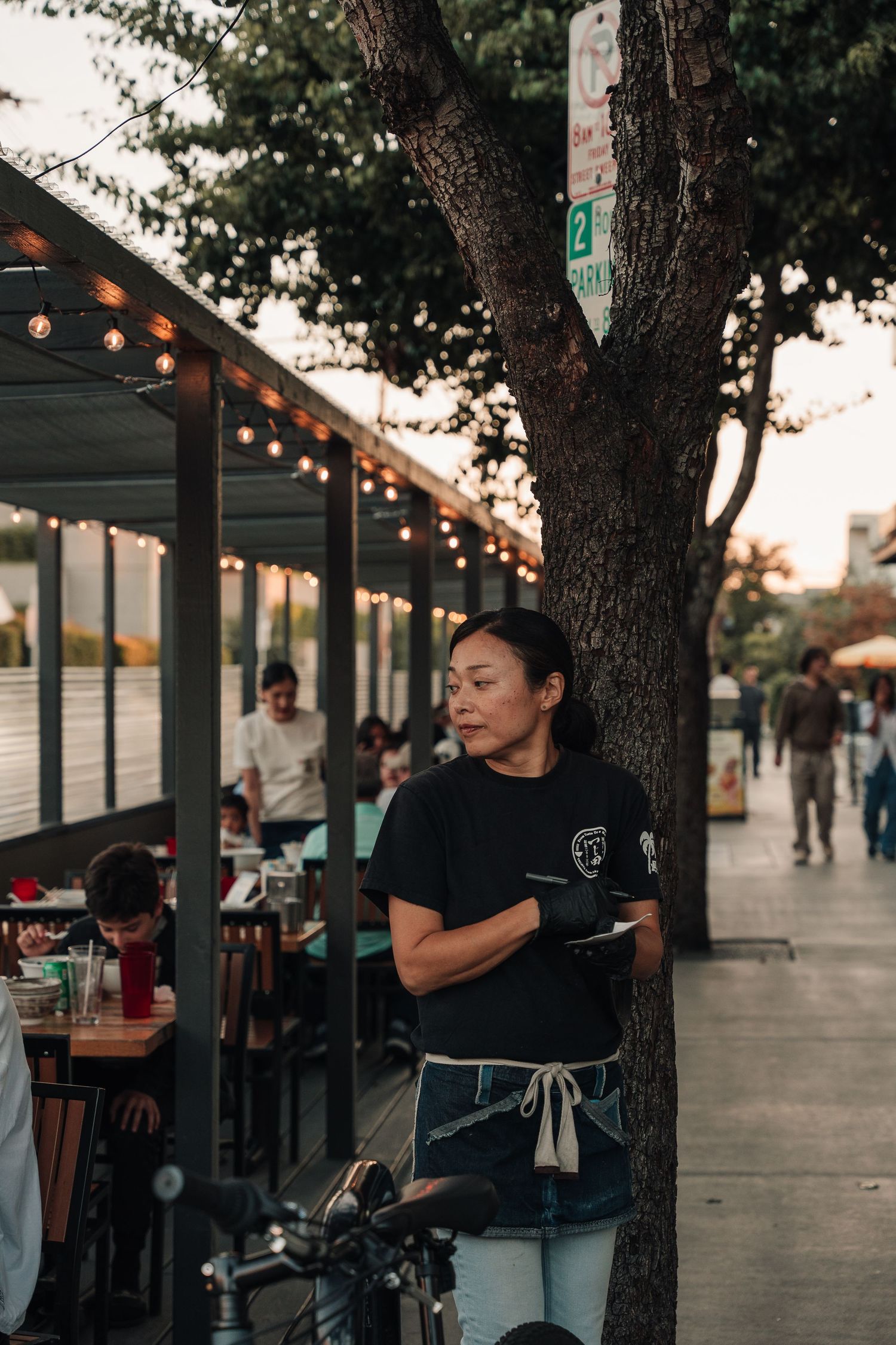 Female server in black uniform and apron standing outside a restaurant patio at sunset.