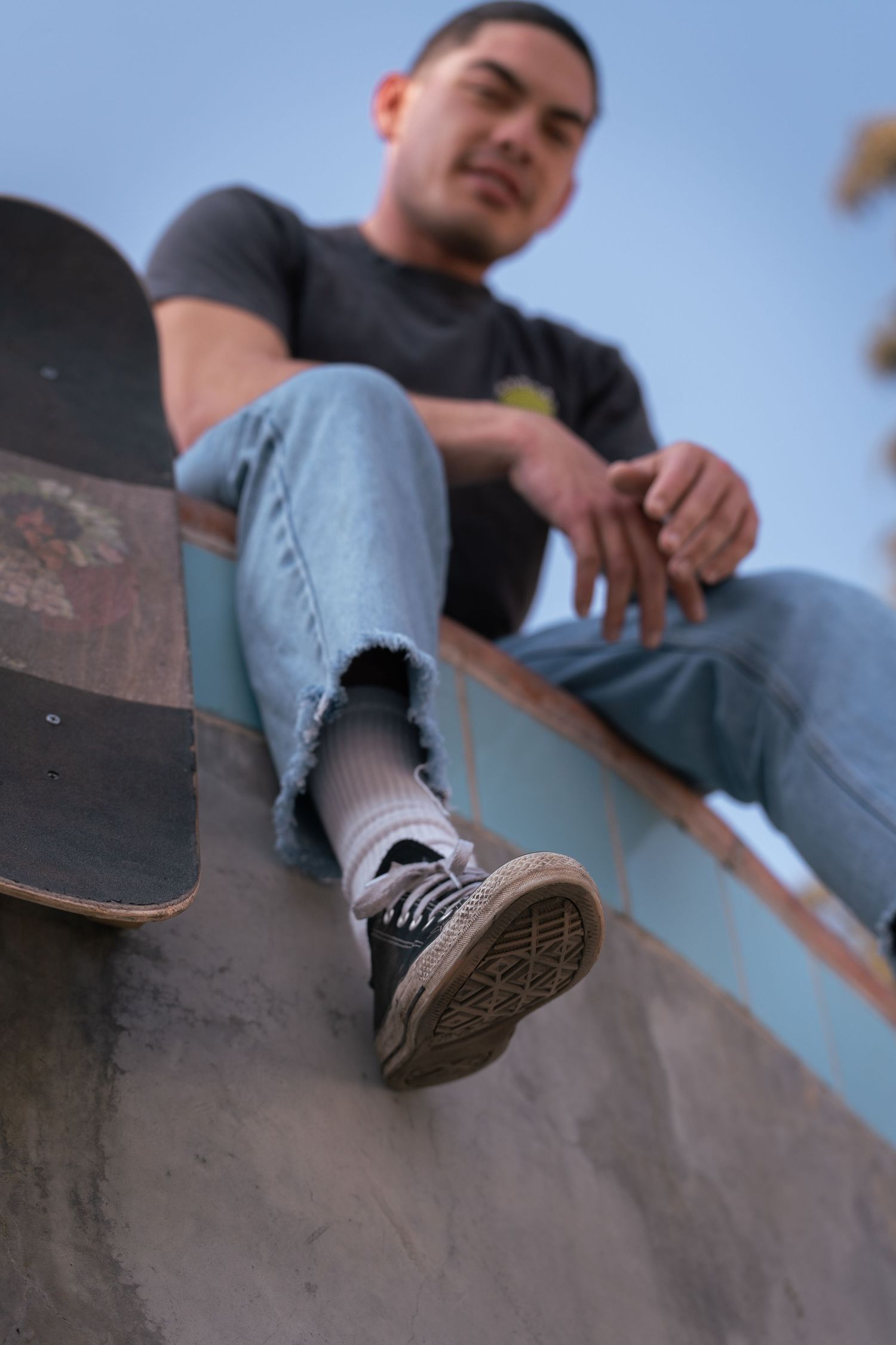 Low angle view of a skateboarder in jeans and sneakers sitting on a concrete ramp with his skateboard.