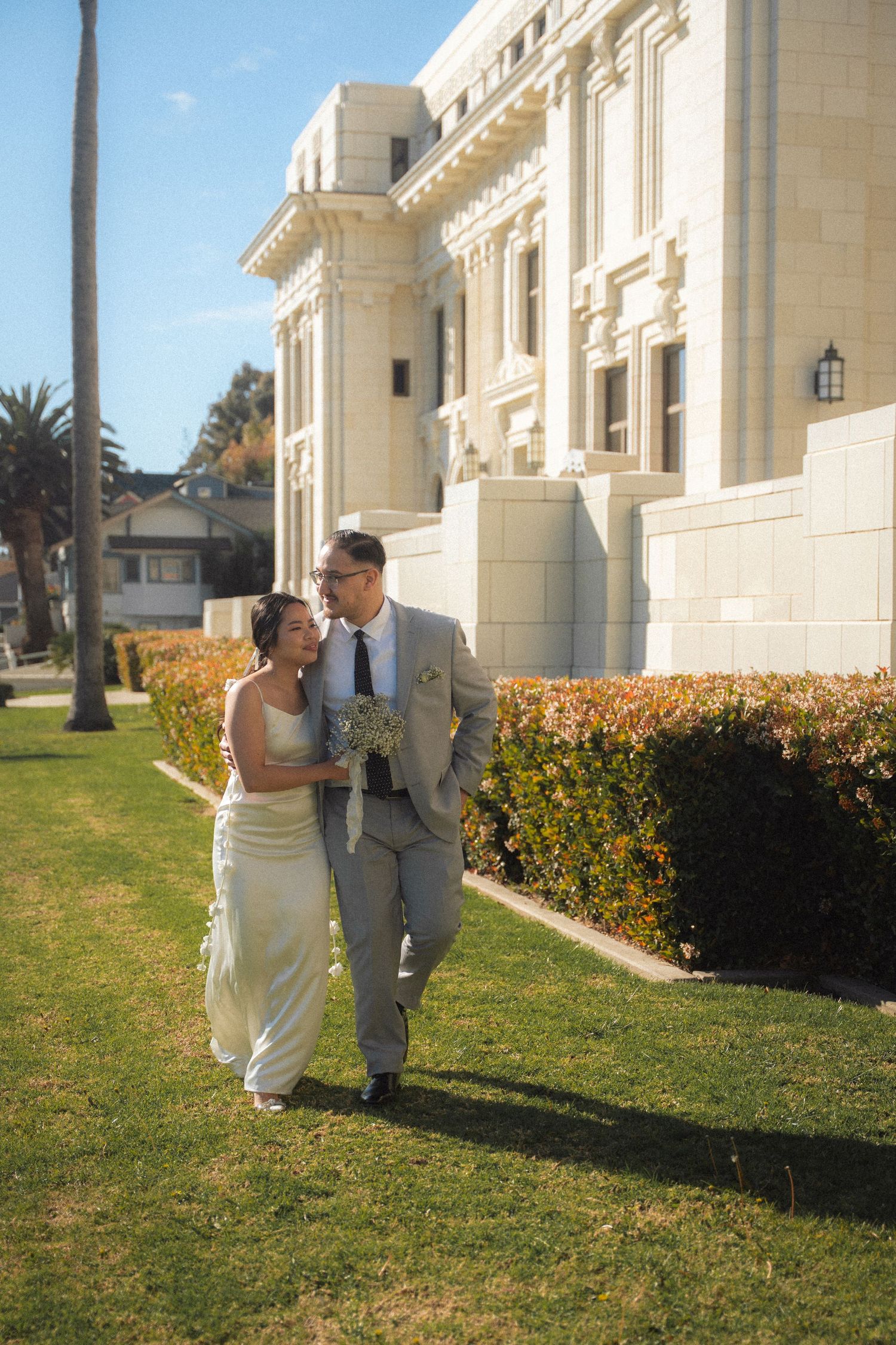 Bride and groom walking together on grass beside grand white neoclassical building on sunny day.