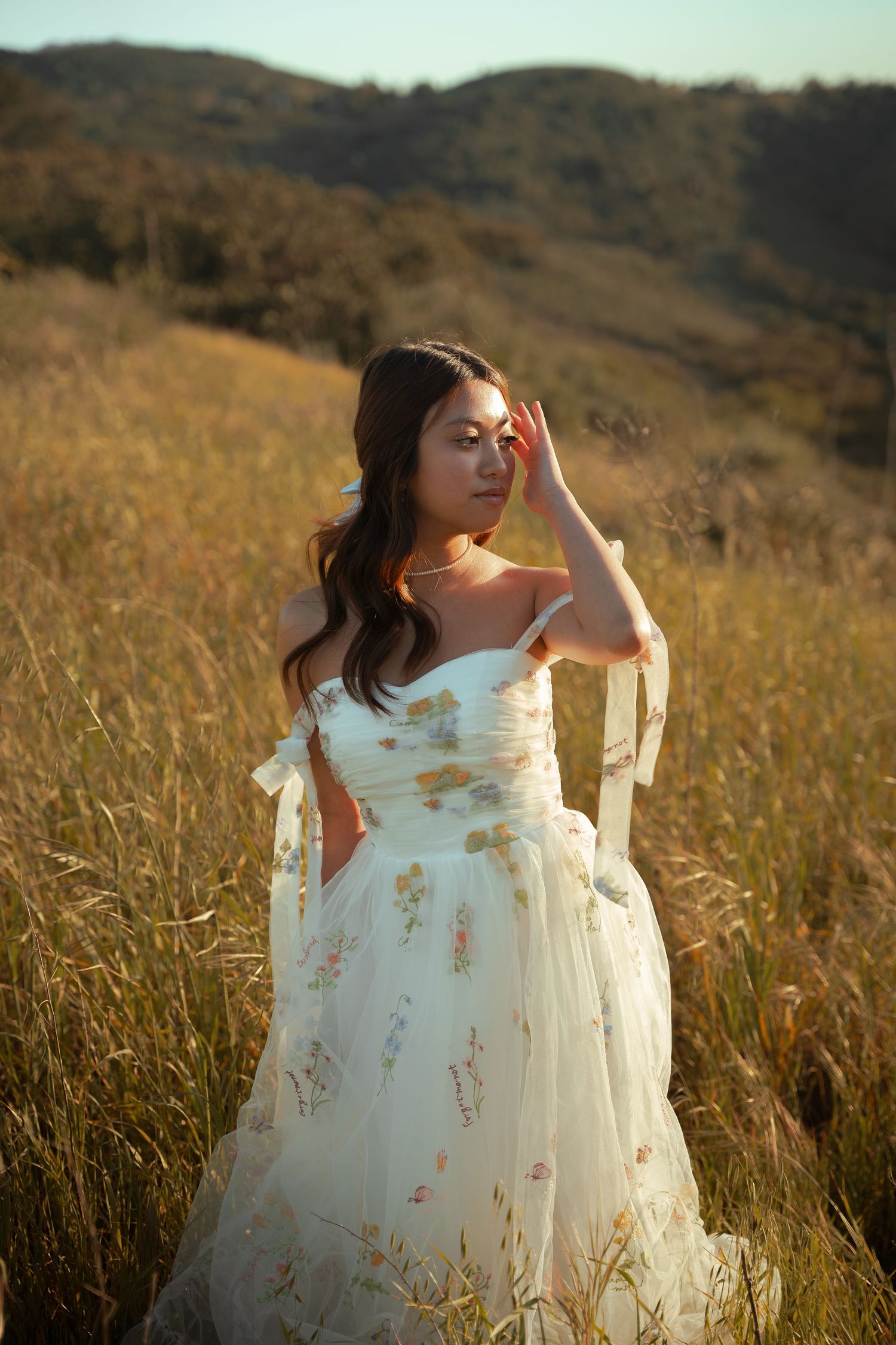 Elegant woman in white floral dress standing in sun-drenched meadow with rolling hills backdrop.