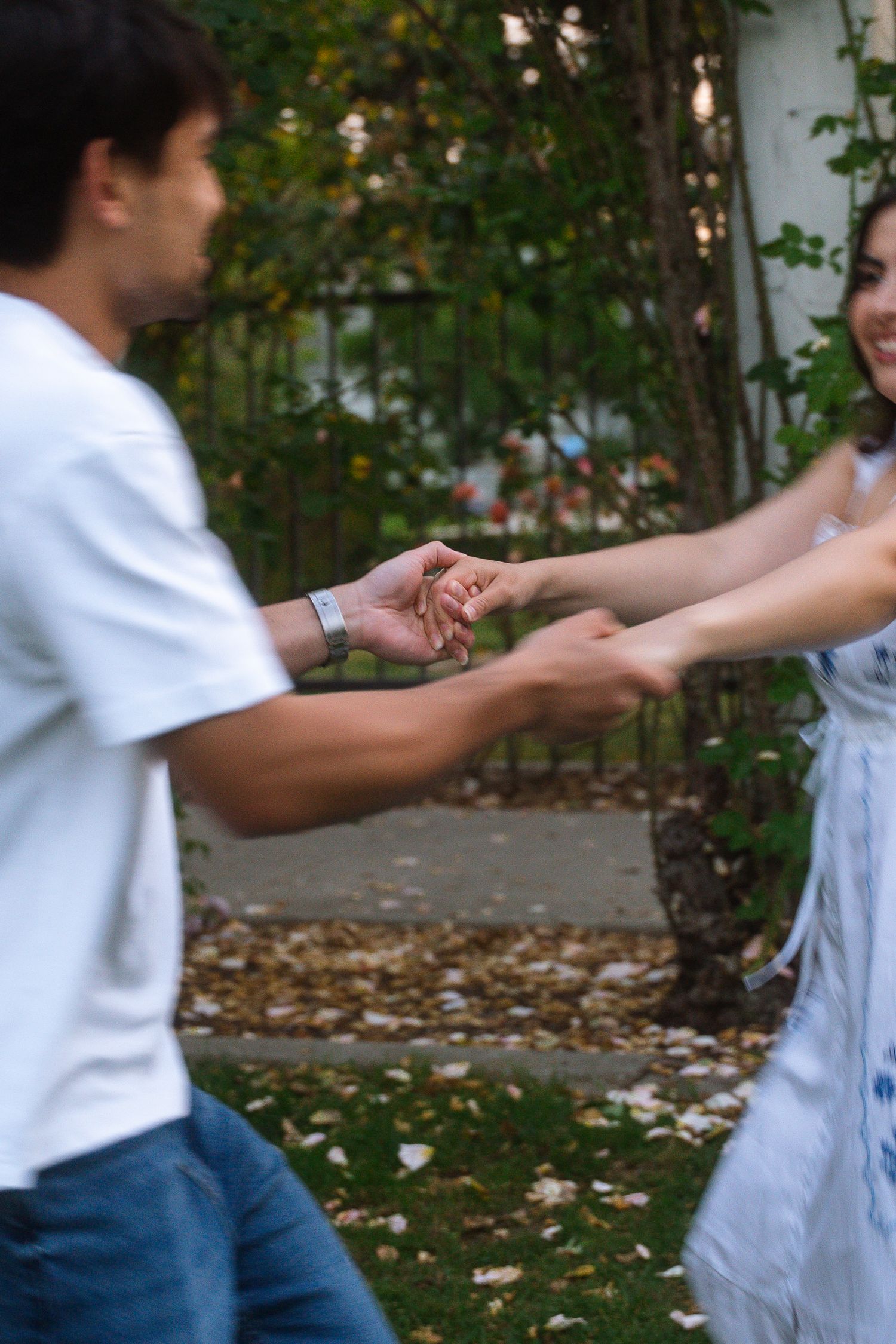 Couple holding hands while spinning outdoors in a garden with autumn leaves on ground.