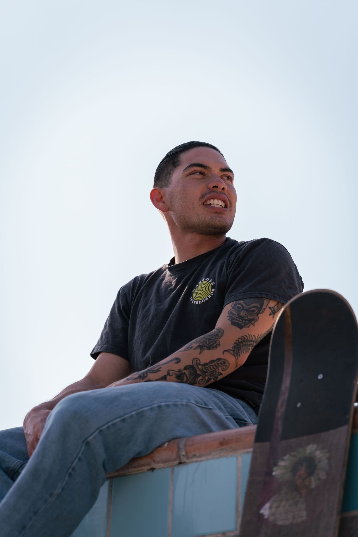 Tattooed young man smiling while sitting on a skate ramp holding his skateboard against a blue sky.