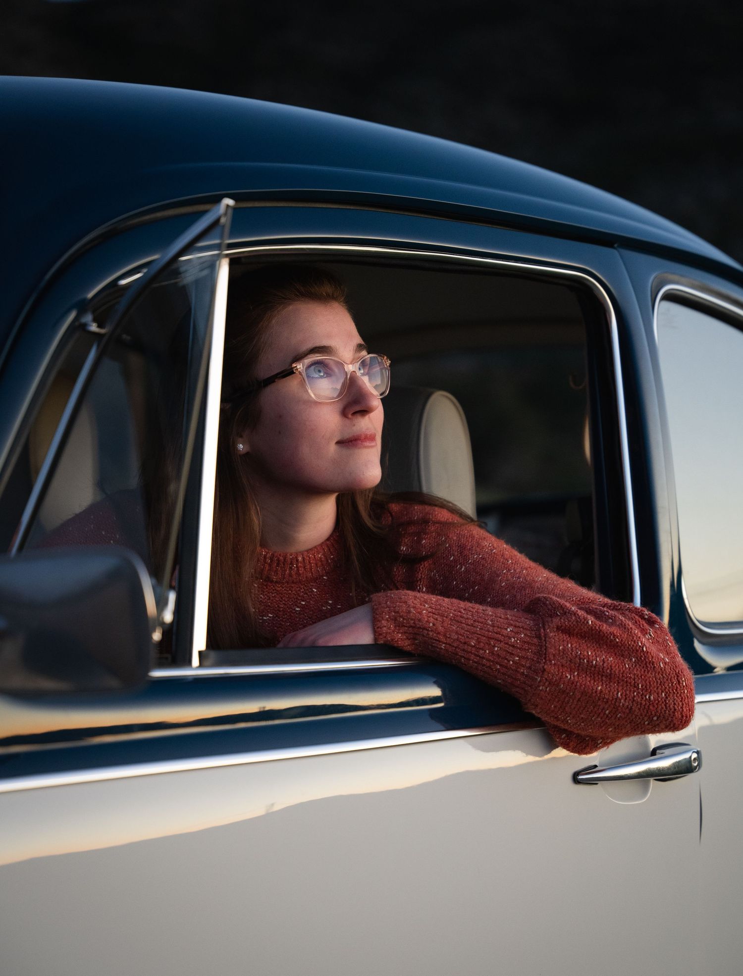 Young woman with glasses in red sweater gazes out window of vintage blue car at sunset.