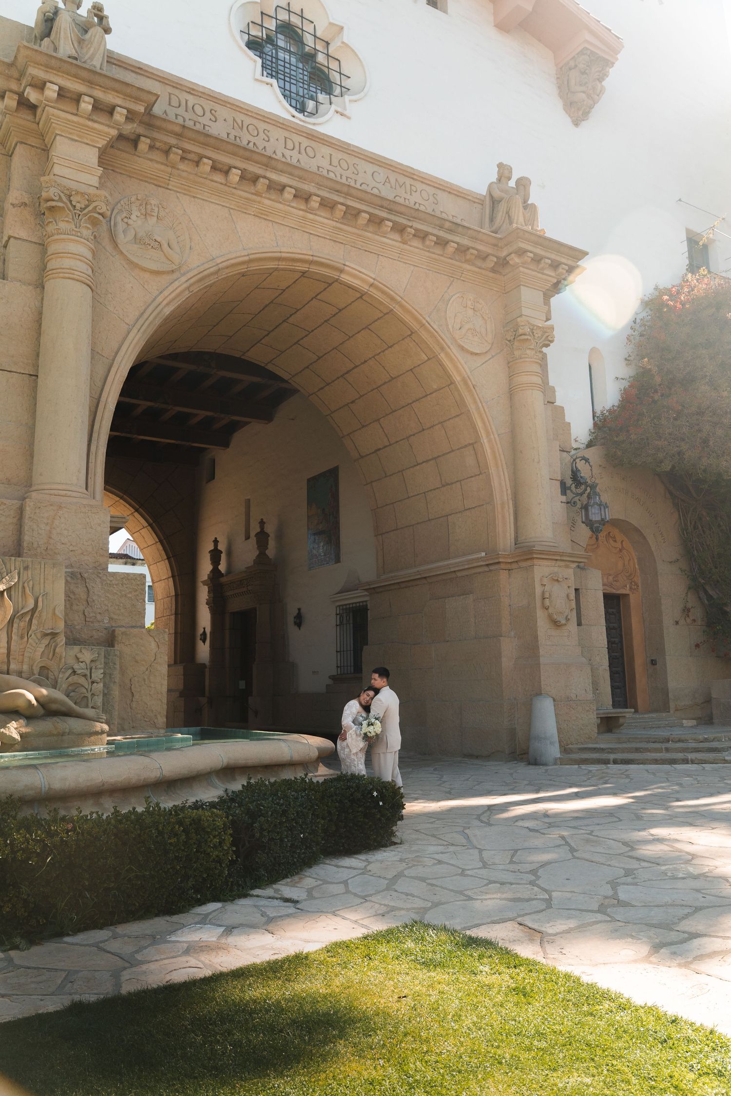 Newlywed couple embracing near ornate fountain outside grand Spanish-style wedding venue arch.