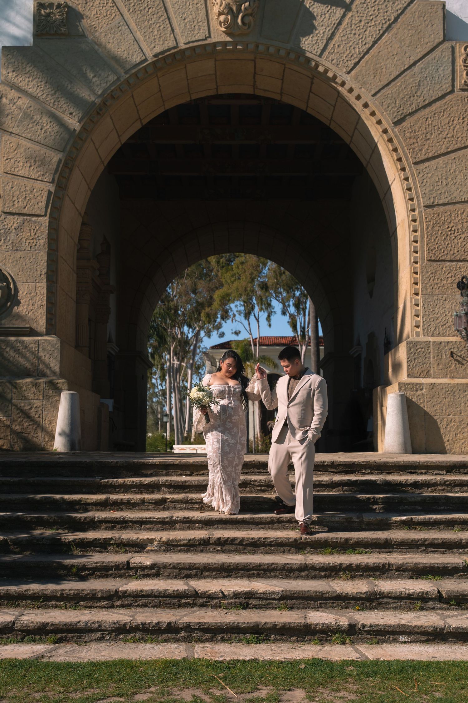 Bride and groom walking down stone steps through decorative arch at historic venue.