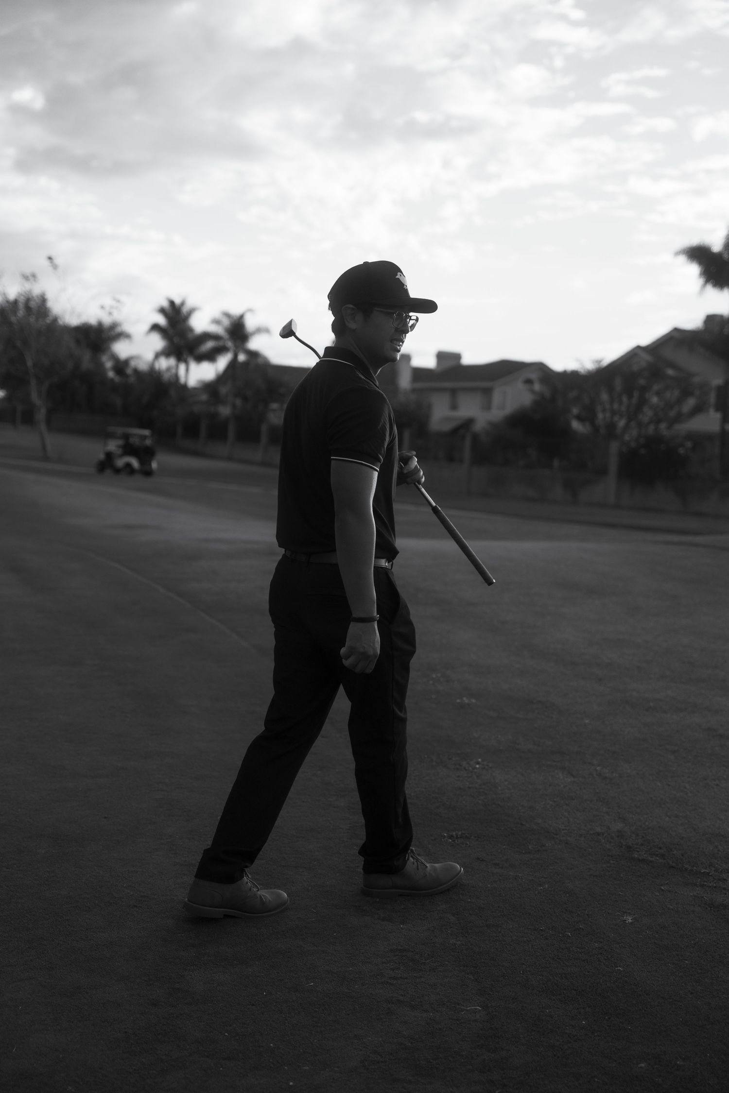 Black and white photo of a golfer carrying a club while walking on a golf course with palm trees and houses in background.