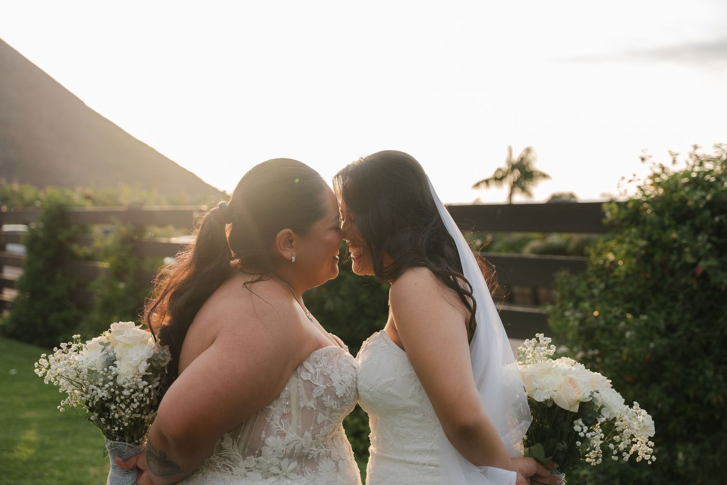 Two brides in white gowns share a tender kiss at sunset with mountains in the background.