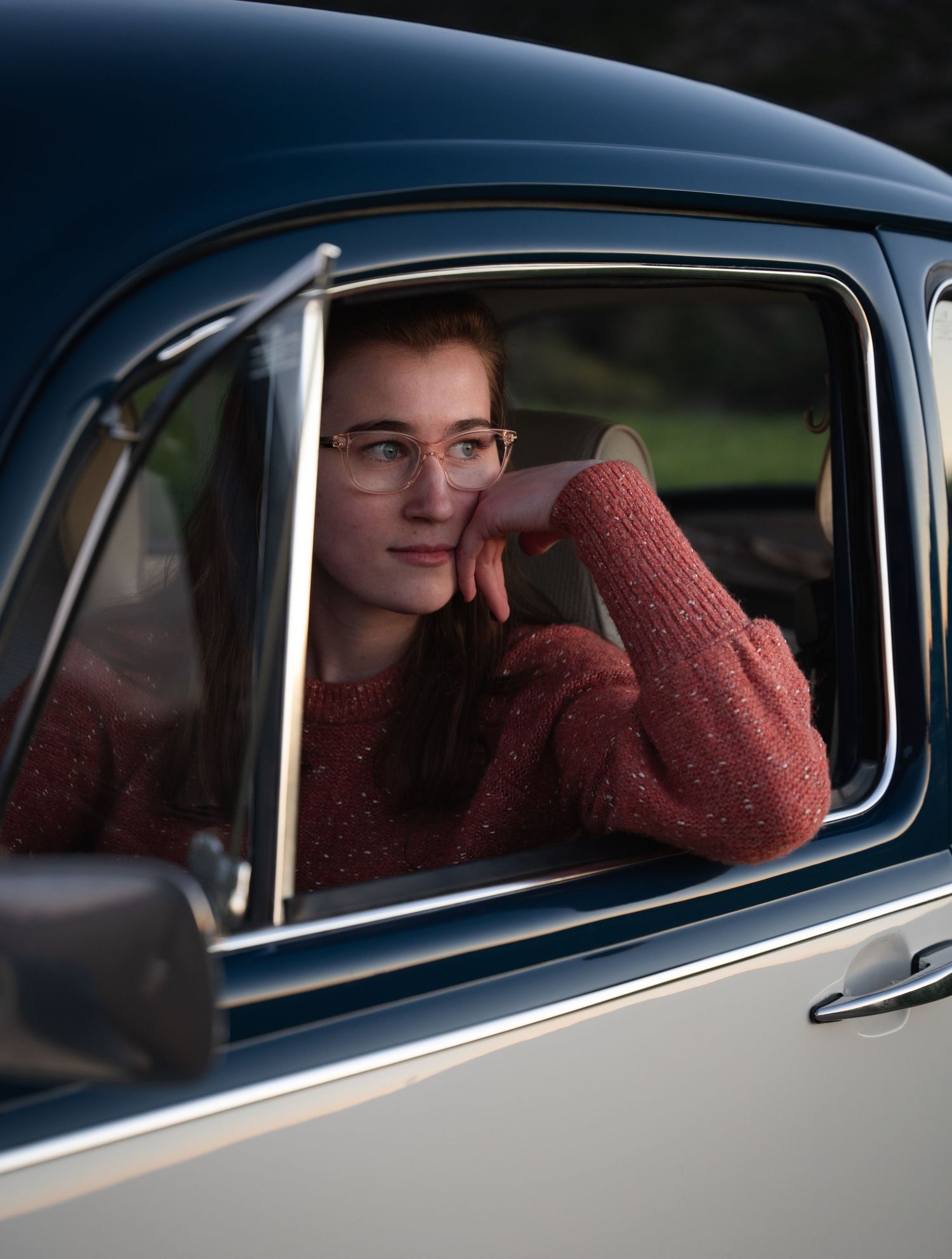 Young woman with glasses in red sweater gazing out vintage blue car window.