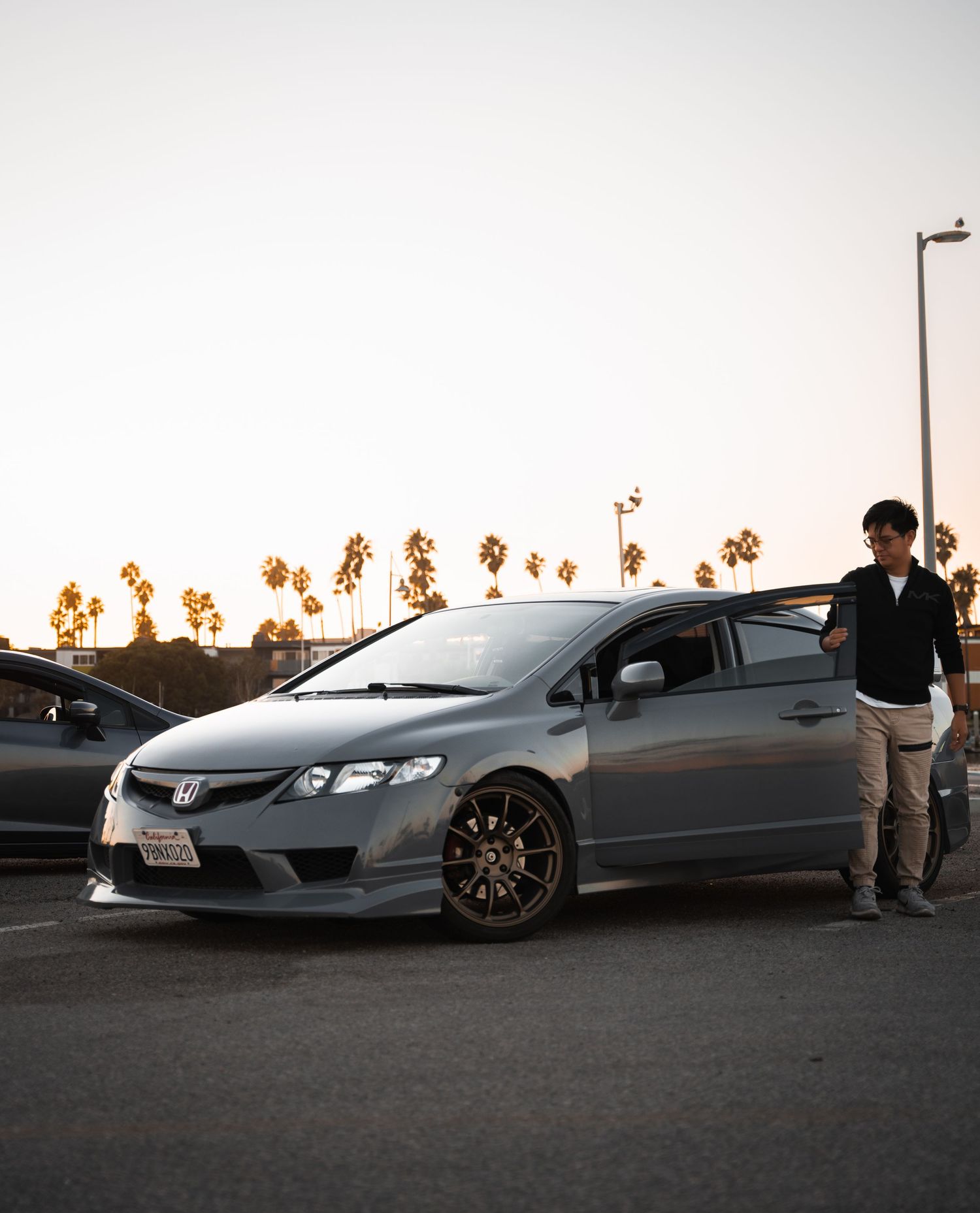 Modified Honda Civic hatchback in silver with bronze wheels parked at sunset near palm trees with owner standing beside it.